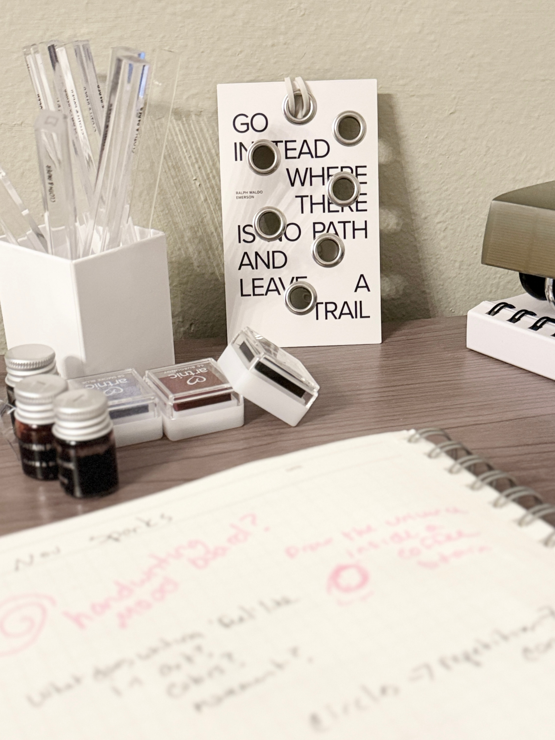 A neatly arranged desk setup featuring a motivational card that reads âGo instead where there is no path and leave a trail,â alongside a spiral notebook, ink vials, and stamp ink pads.