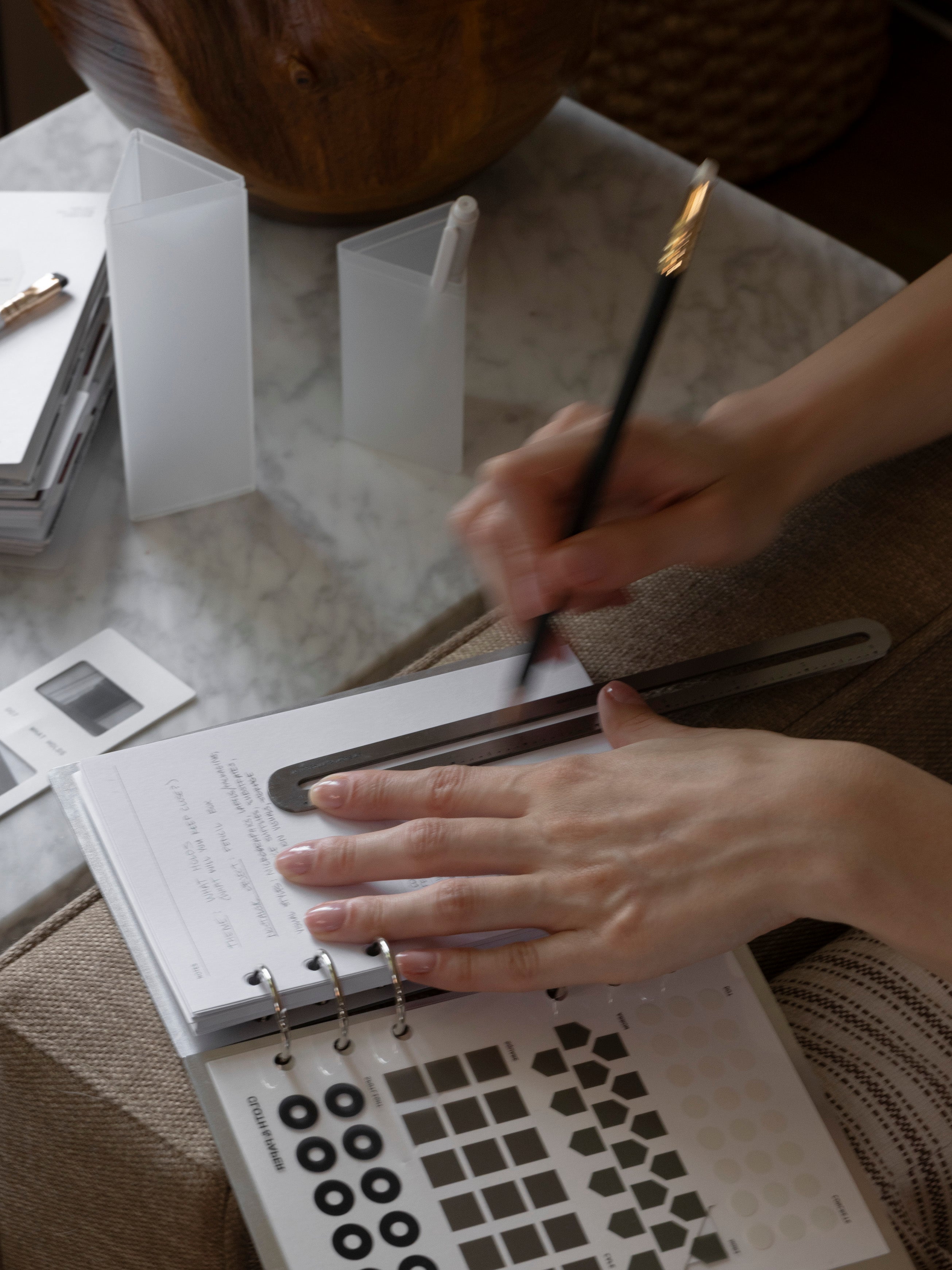 Hands writing in an open Personal Wide Clad Cover Set on a woven surface, with a frosted Pencil Box, punched sticker sheet, and additional planner accessories visible nearby