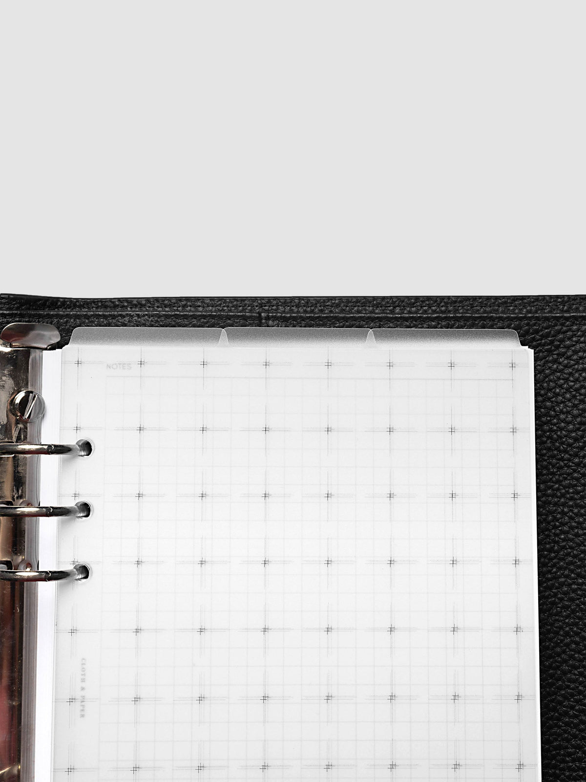 Grid Top Tab Dividers | Ultra-Thin, A5 6-Ring Punch, cropped overhead view of the top of a black leather ring bound planner with silver rings, three frosted tabs staggered above the grid printed page