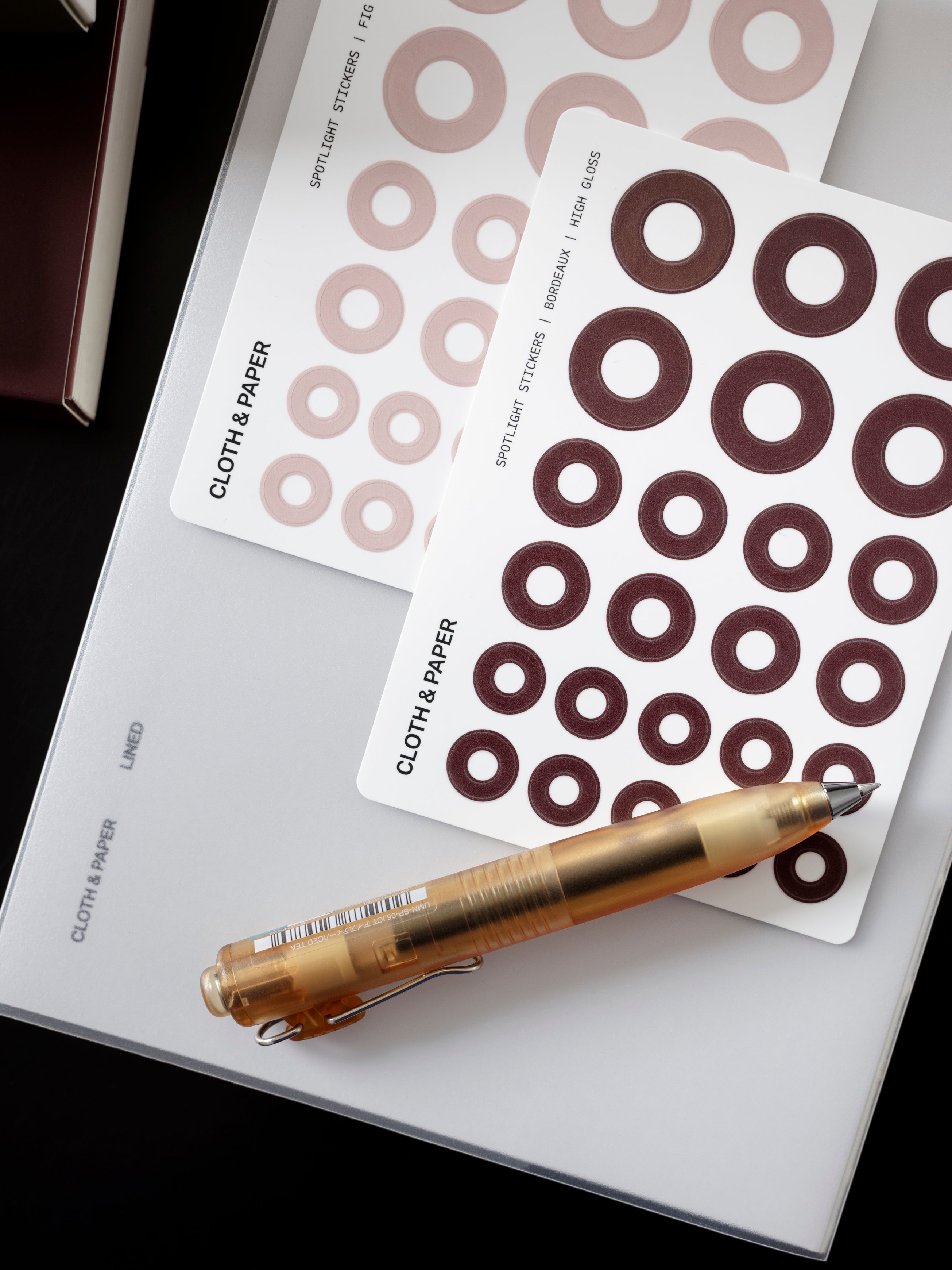 One sticker sheet each of Fig Milk and Bordeaux Spotlight Stickers | High Gloss on top of a notebook with a pen resting across them.
