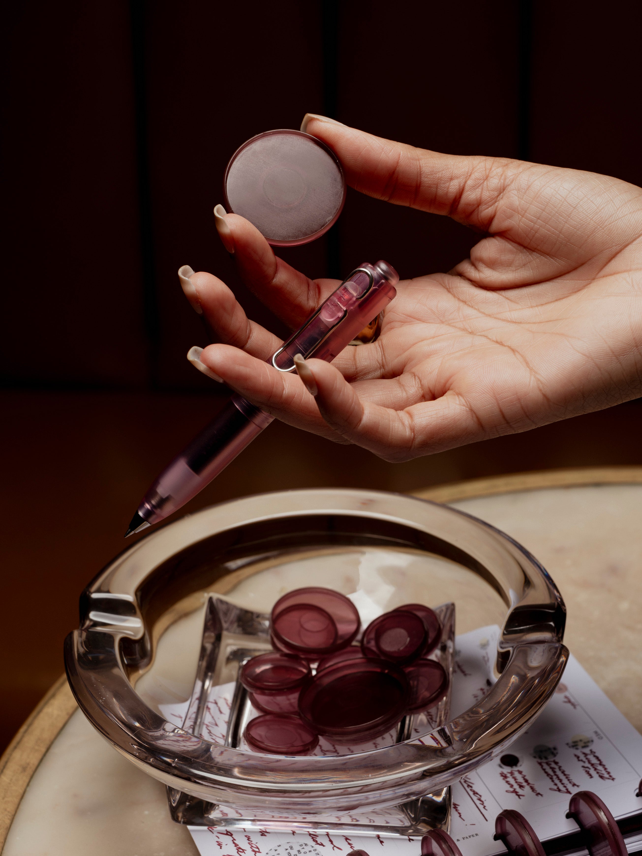 Hand holding a Transparent Planner Disc in the color Bordeaux alongside a matching color pen with objects on the table below.