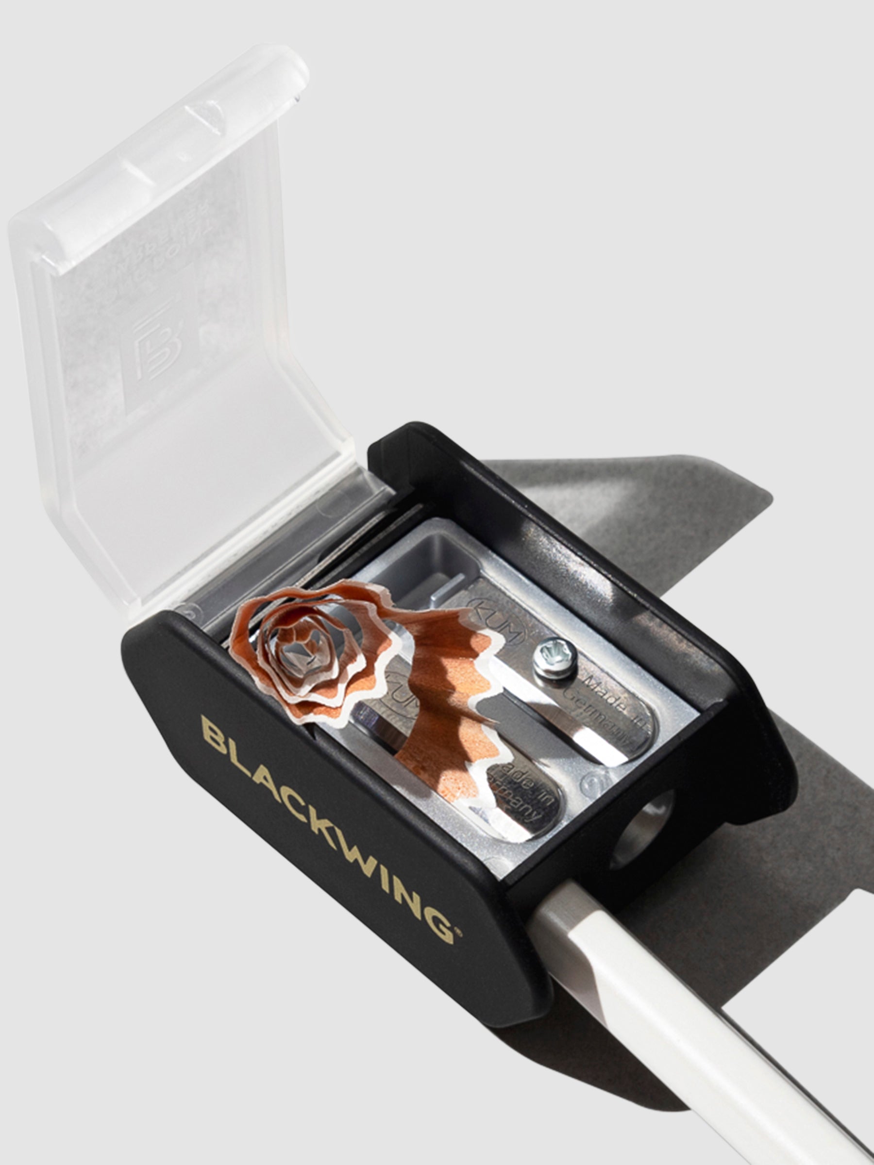 The image shows the blackwing long point pencil sharpener. It features perfectly curled pencil shavings as a white pencil is sharpened.