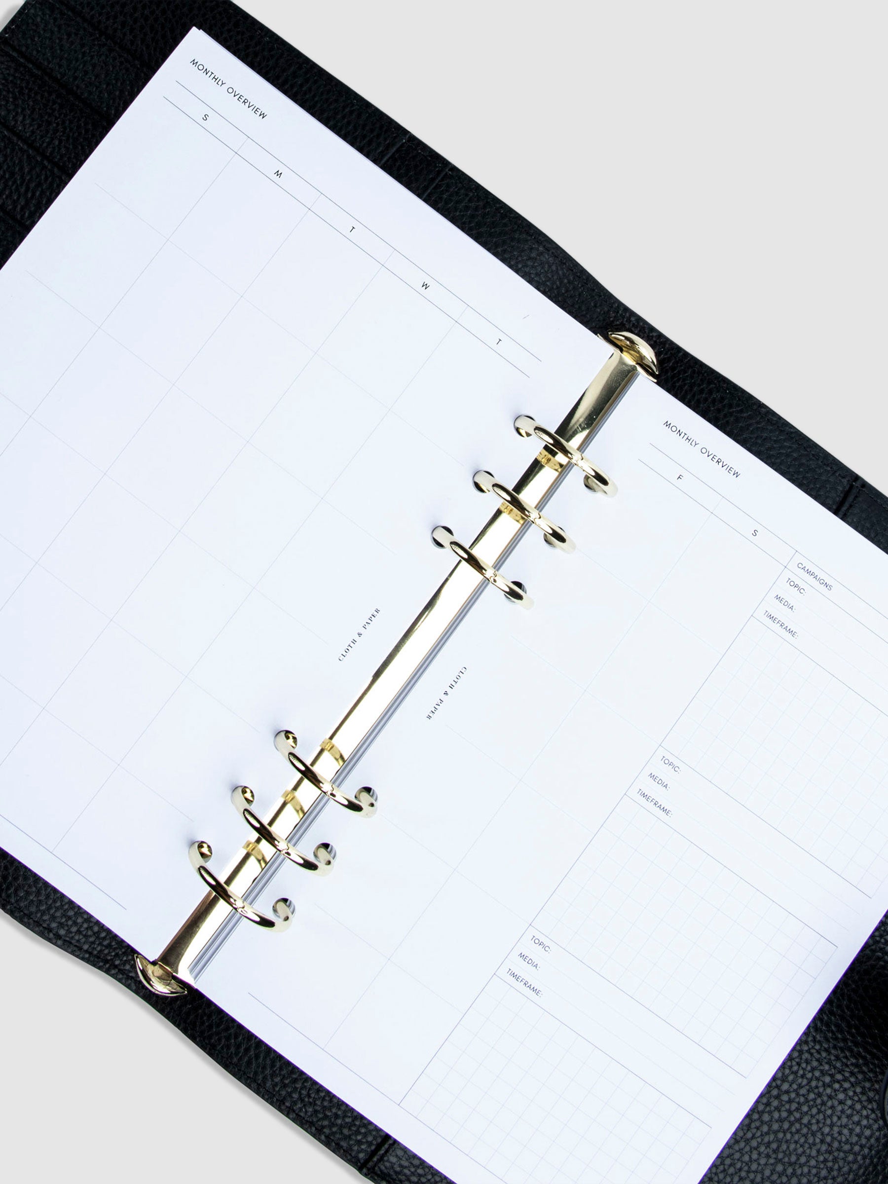 Creative Planning Insert Bundle, Cloth and Paper. Inserts in use inside a black leather agenda with gold rings tilted to the right. Insert spread shows the Monthly Overview.