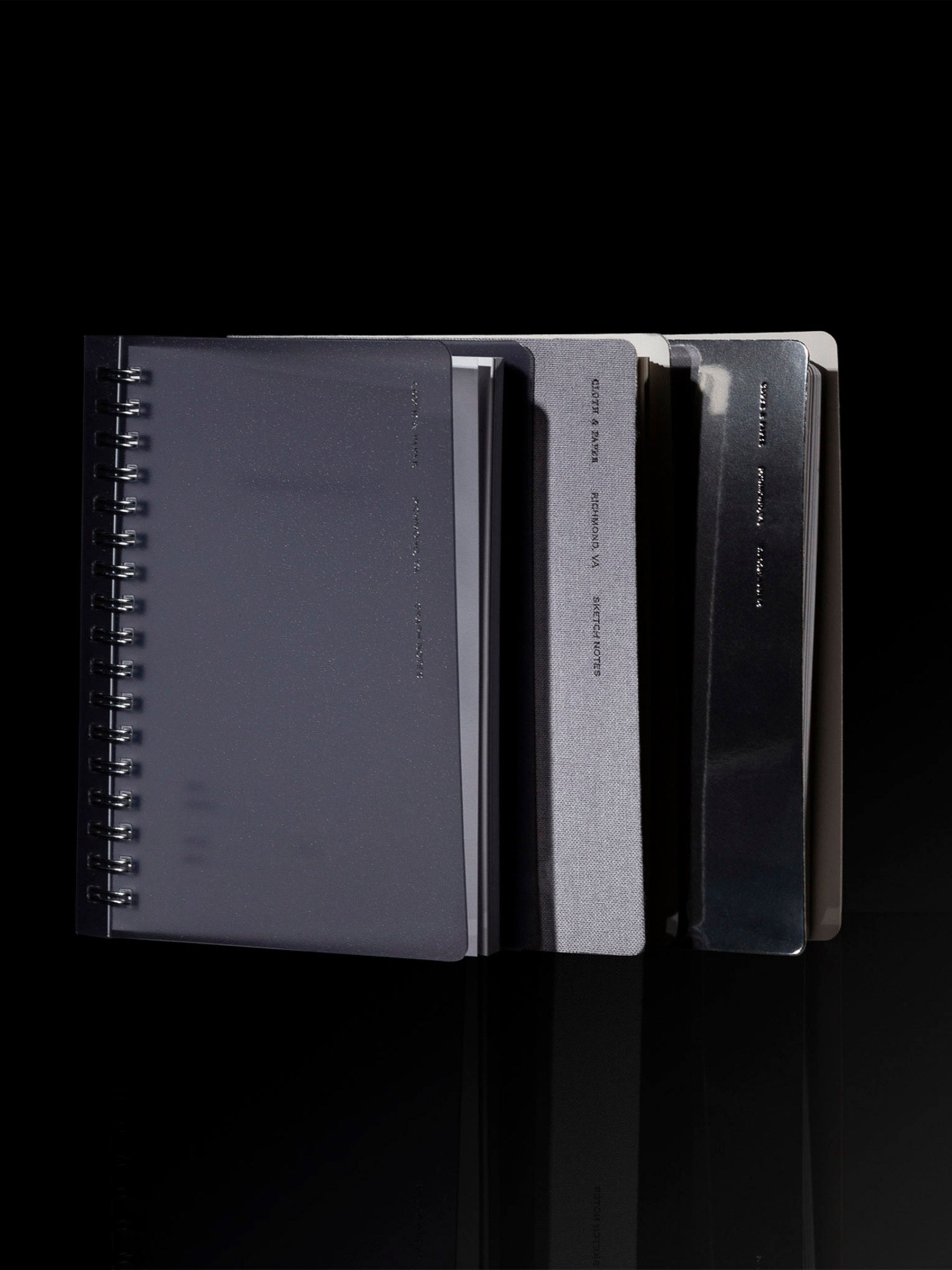 Collection of three notebooks, silver, tinted plastic, and linen, displayed on a black background in dramatic strong lighting.