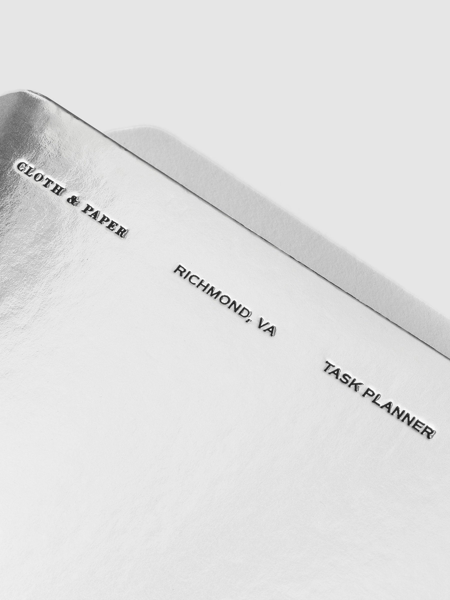 Closeup of silver notebook. Embossed text reads "Cloth & Paper | Richmond, VA | Task Planner"