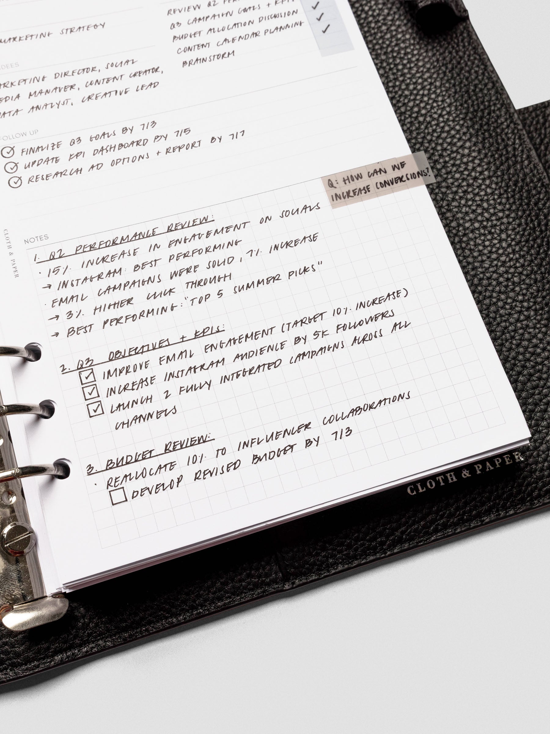 Close-up of a meeting notes insert inside a black leather planner, filled with handwritten agenda points, follow-up tasks, and gridded notes.