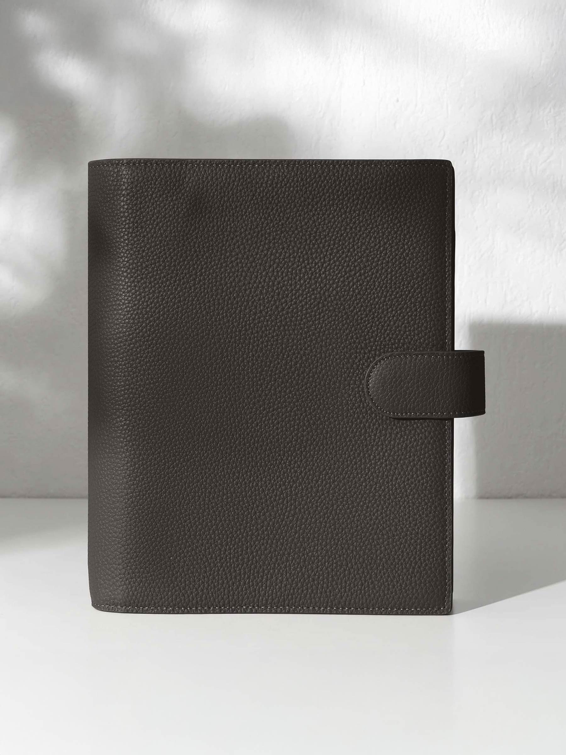 A closed cafenoir leather planner with a closure displayed against a shadowed background.