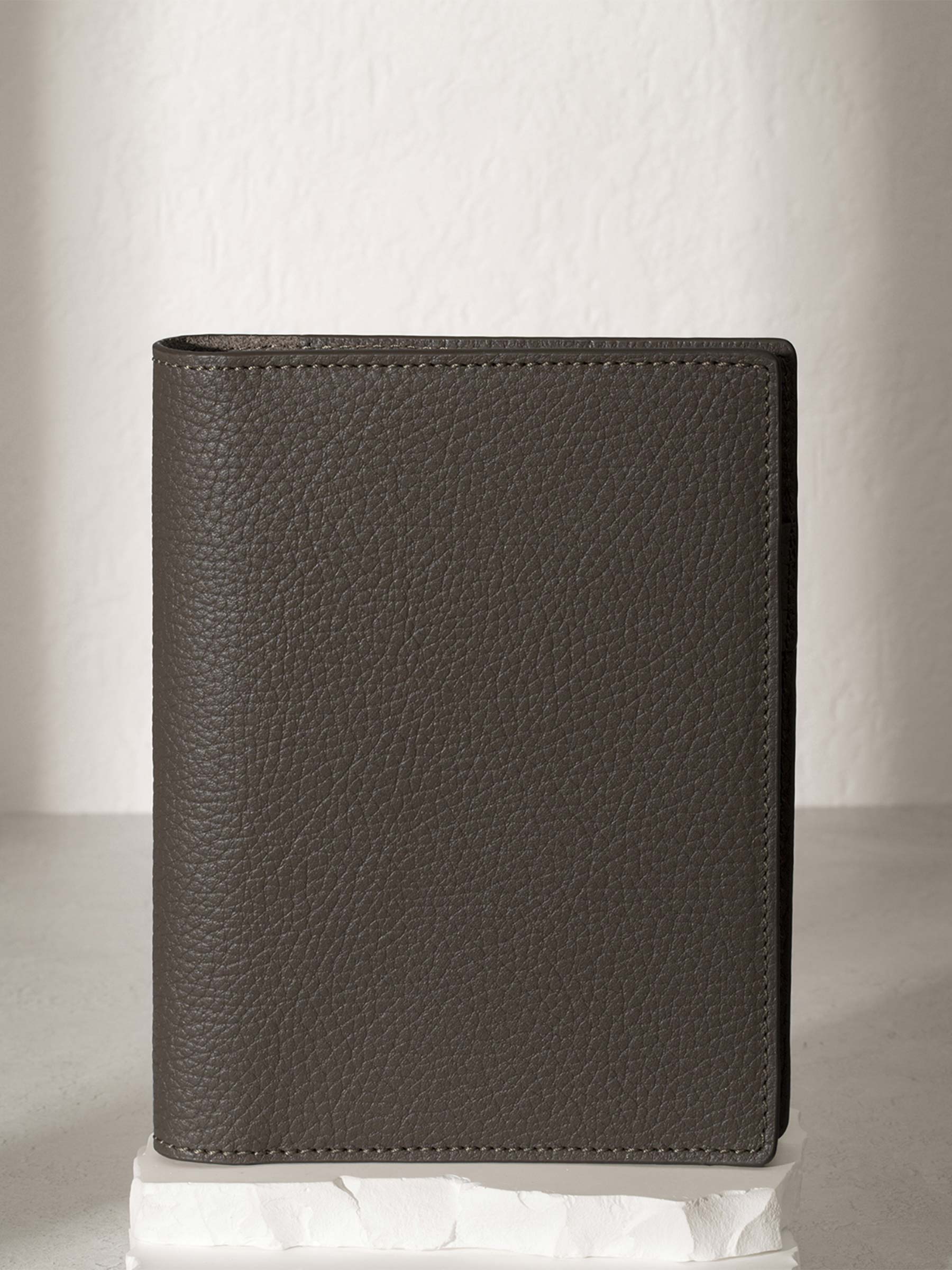 CP Petite Leon folio displayed on a white stone pedestal. The background is a natural textured off-white material, and a spotlight behind the folio highlights its placement.