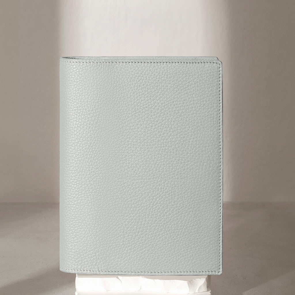 Veleta folio displayed on a white stone pedestal. The background is a natural textured off-white material, and a spotlight behind the folio highlights its placement.