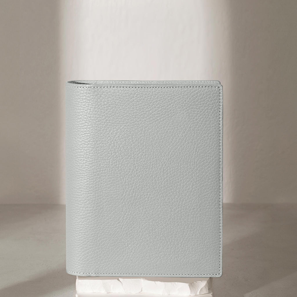 Heirloom Leather Folio, Small, Cloth and Paper. Veleta folio displayed on a white stone pedestal. The background is a natural textured off-white material, and a spotlight behind the folio highlights its placement.