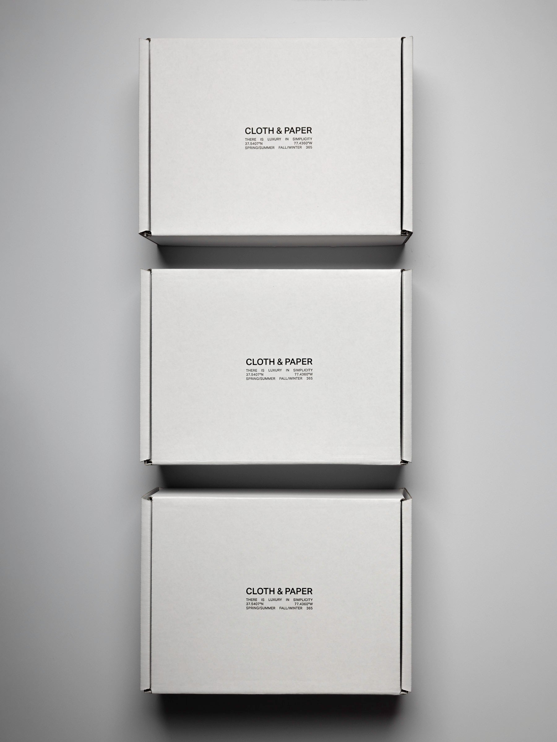 Three white, sealed boxes, viewed from the top down on a neutral background. In black lettering on the top reads "Cloth & Paper. There is luxury in simplicity".