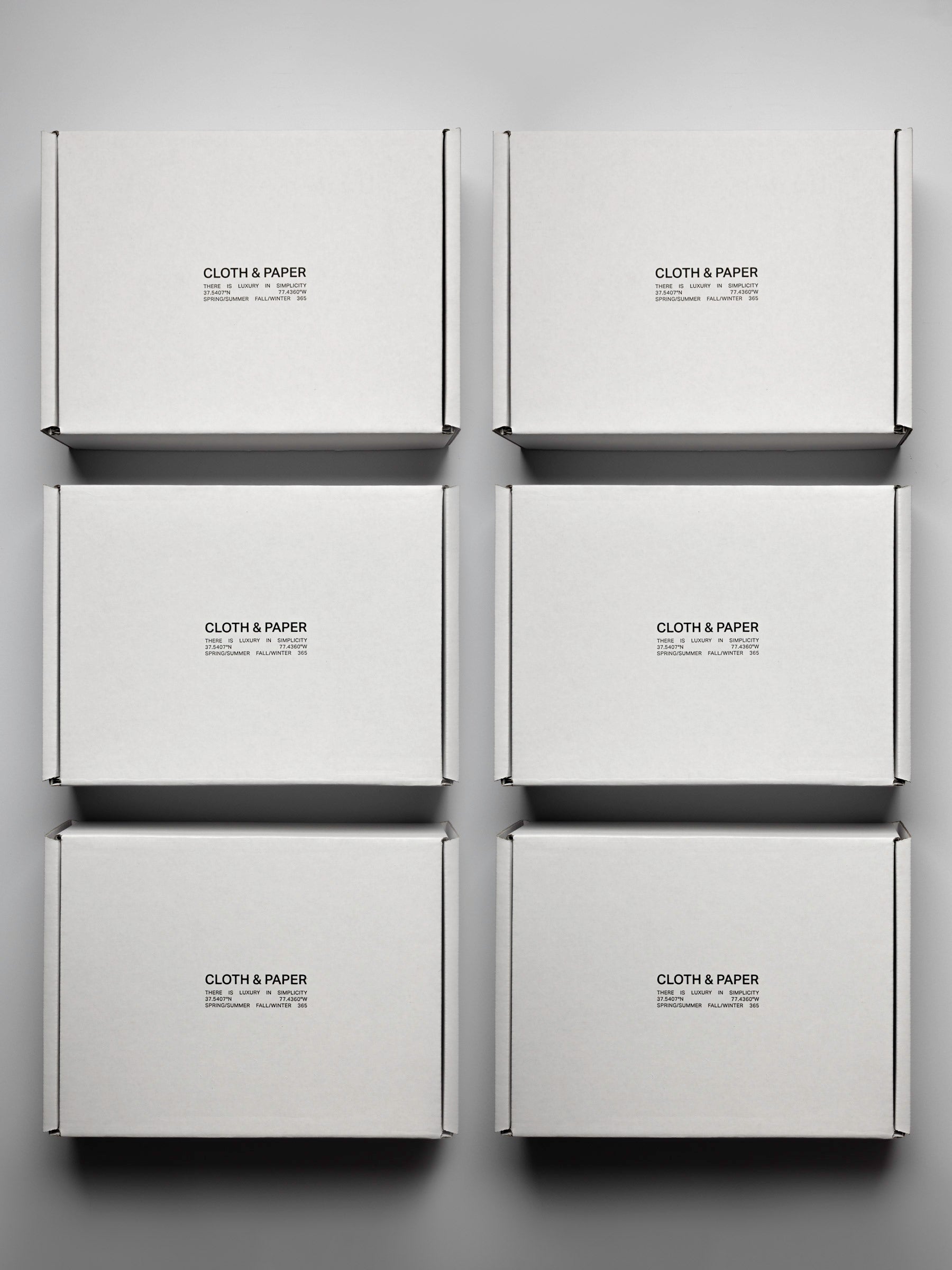 Six white, sealed boxes, viewed from the top down on a neutral background. In black lettering on the top reads "Cloth & Paper. There is luxury in simplicity".