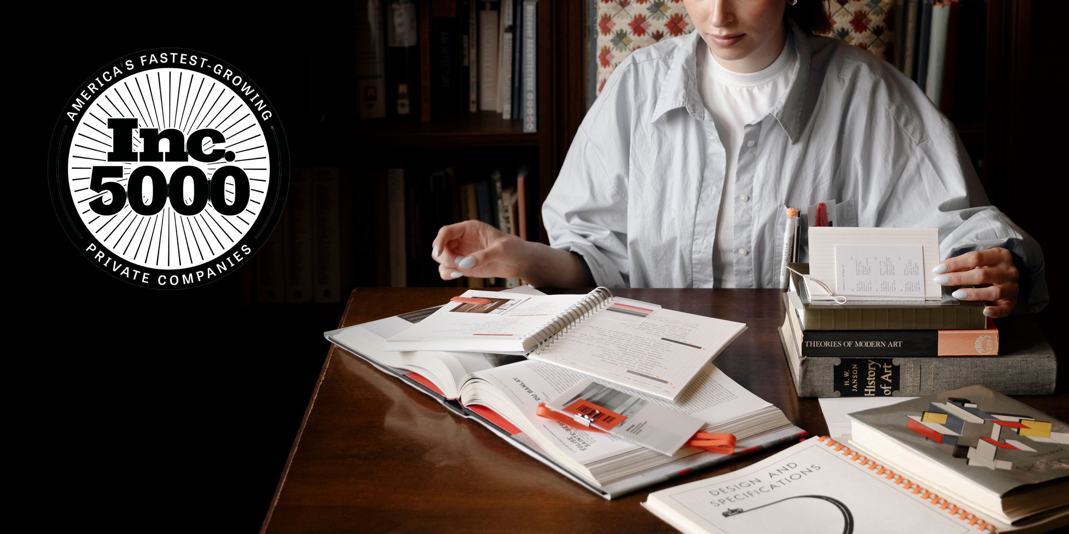 A woman is shown browsing through a pile of several books and notebooks displayed on a desk. In the upper left corner is a logo for Inc. 5000 as Cloth & Paper was recognized for being one of America's Fastest-Growing Private Companies.