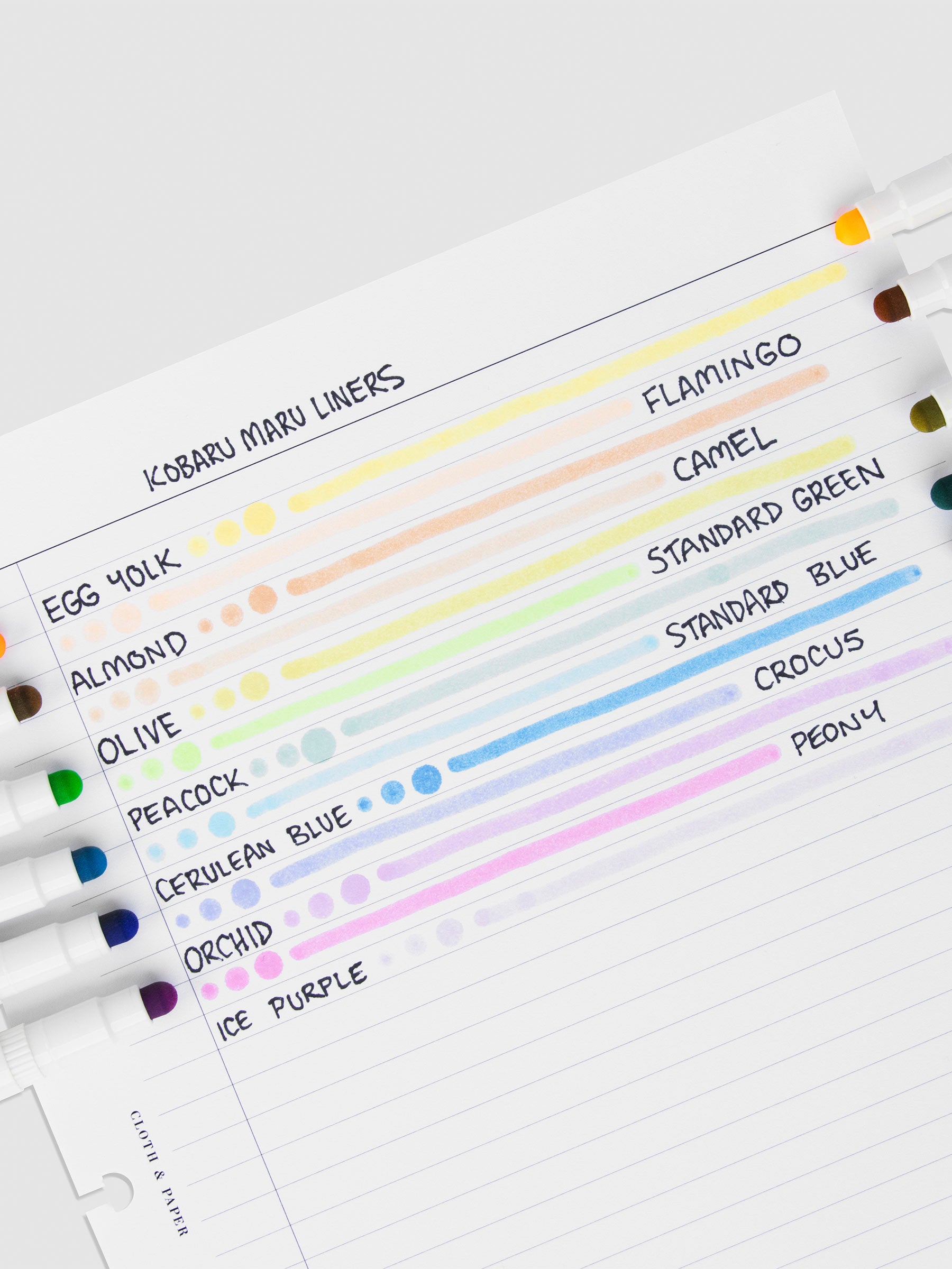 Pen test sheet showing examples of Egg Yolk, Flamingo, Almond, Camel, Olive, Standard Green, Peacock, Standard Blue, Cerulean Blue, Crocus, Orchid, Peony, Tourmaline, and Ice Purple ink. The pens are resting on the pen test sheet.