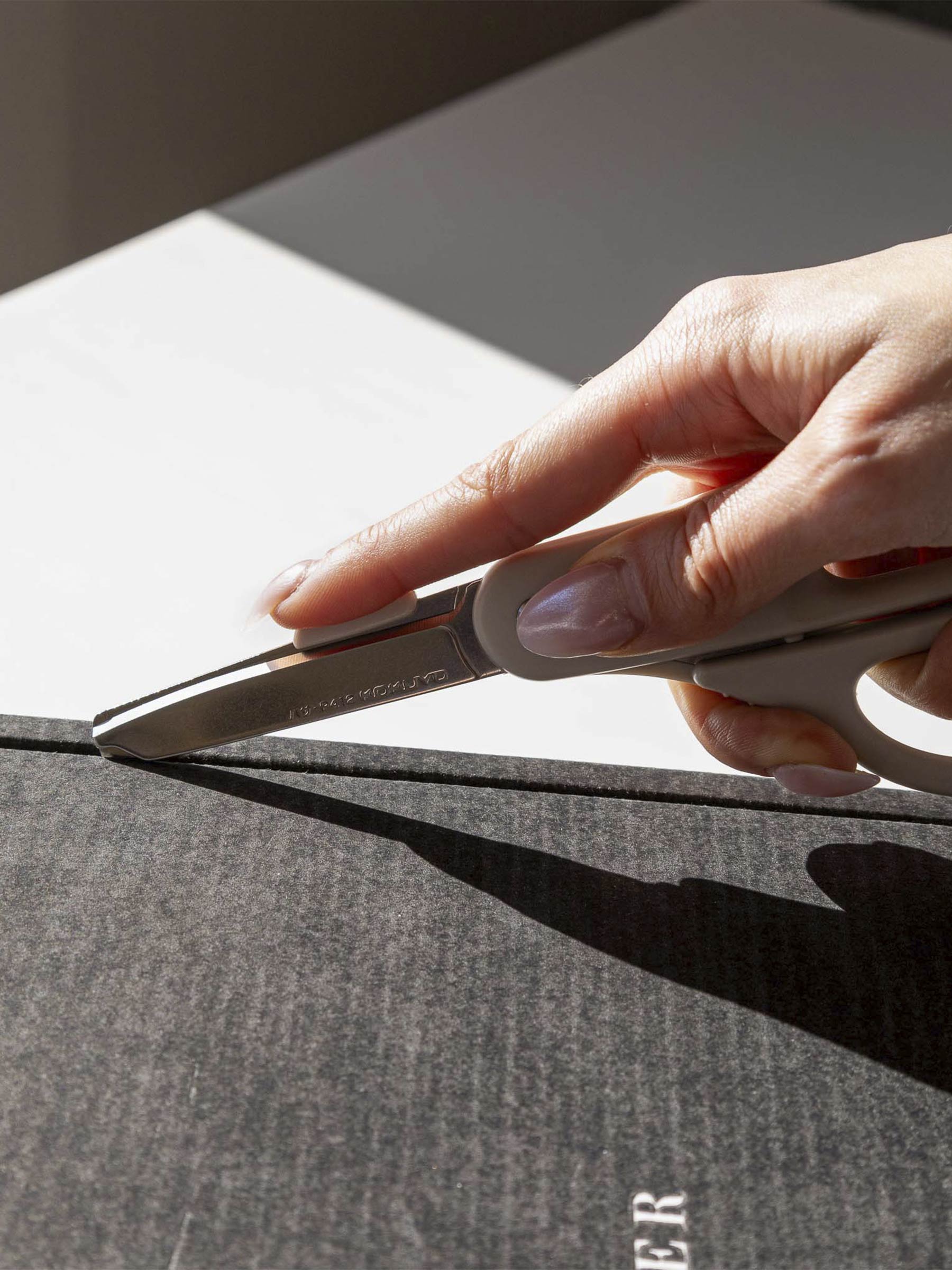 A pale hand shown using a pair of scissors in cutting mode to unseal a Cloth and Paper box.