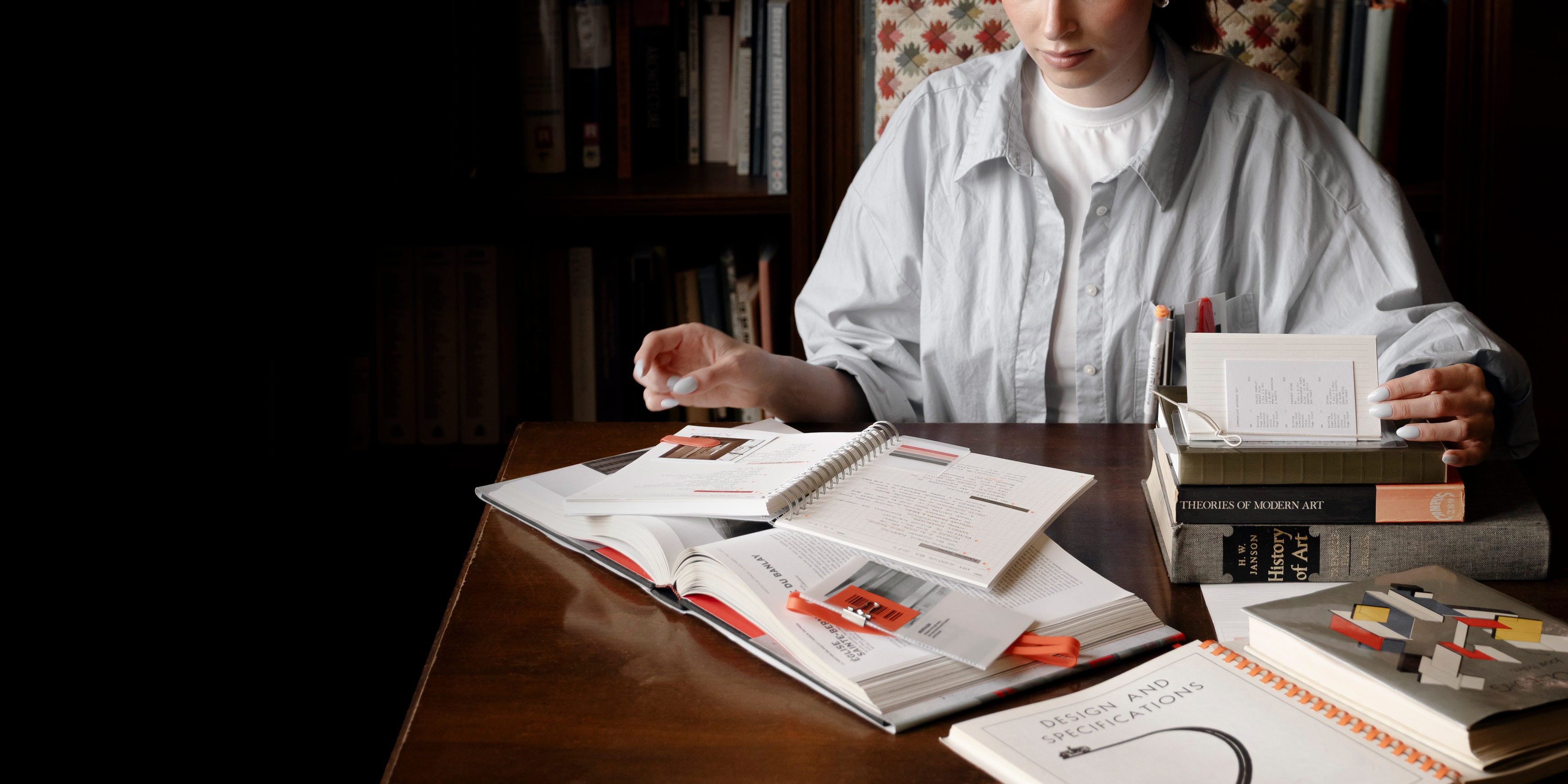 A woman is shown browsing through a pile of several books and notebooks displayed on a desk.