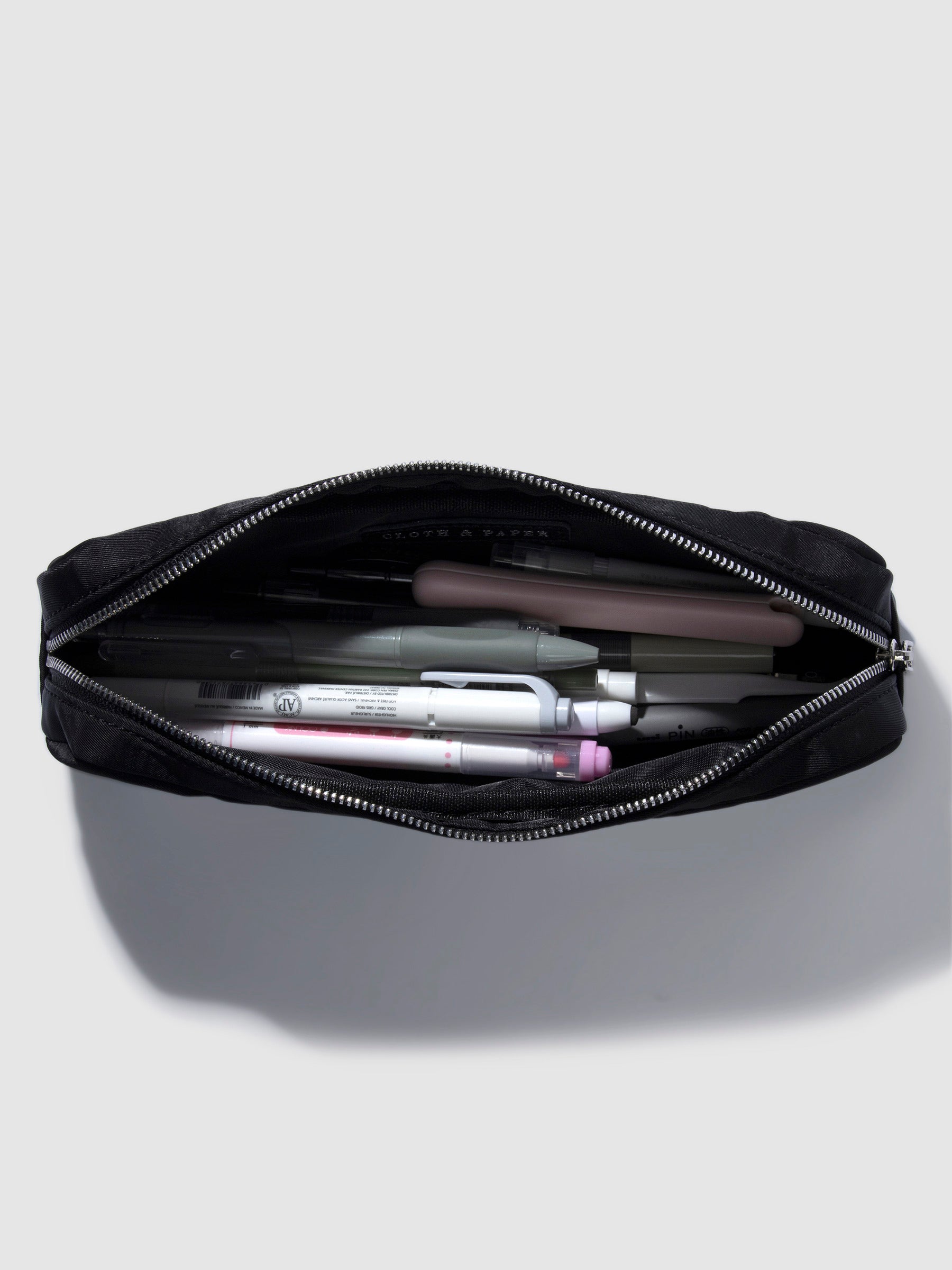 Pencil pouch displayed unzipped with a collection of pens and accessories stored inside. Color featured is black.
