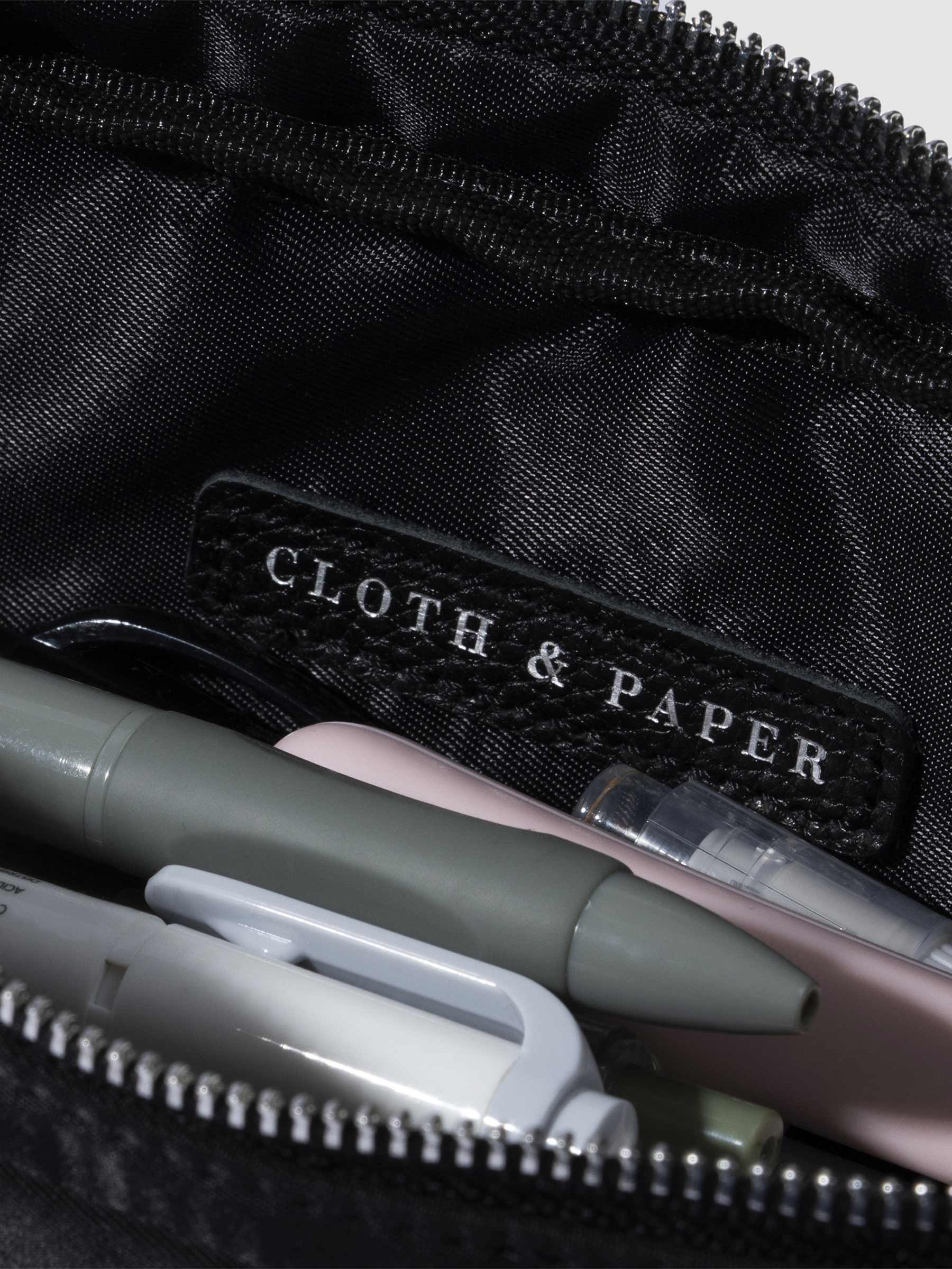 Closeup of Cloth and Paper logo inside of pencil pouch. A selection of pens are stored inside the pouch. Color featured is black.