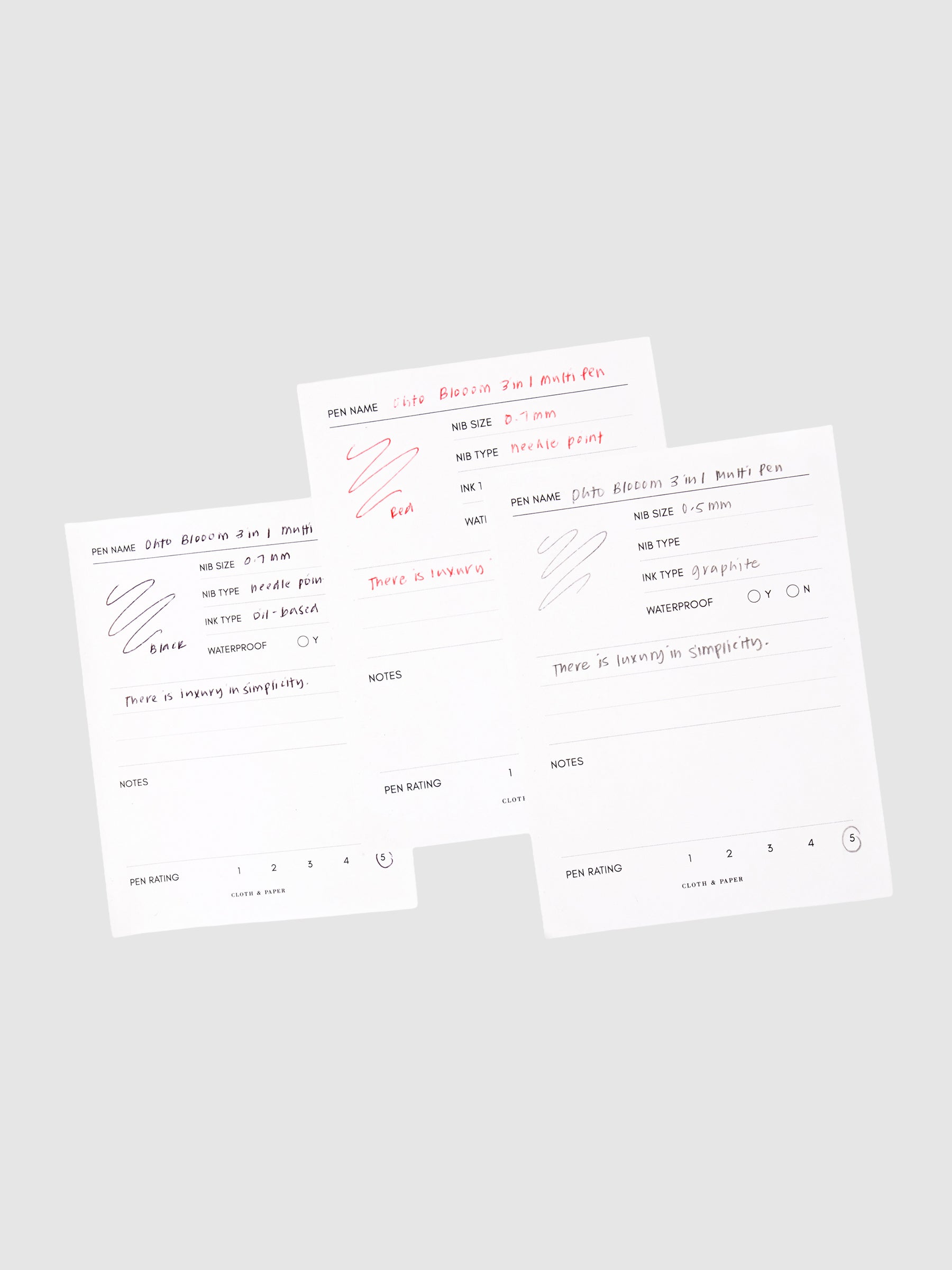 Ohto Bloom 3 in 1 Multi Pen, Cloth and Paper. 3 pen test sheets with writing samples for black ink, red ink, and mechanical pencil layered on top of each other against a neutral background.