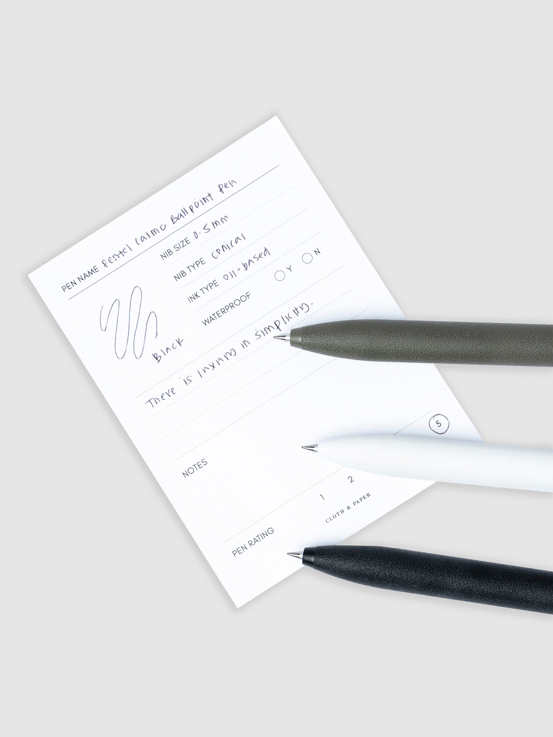 All three color options of the pen resting on a pen test sheet displaying a writing sample detailing the pen's specs.