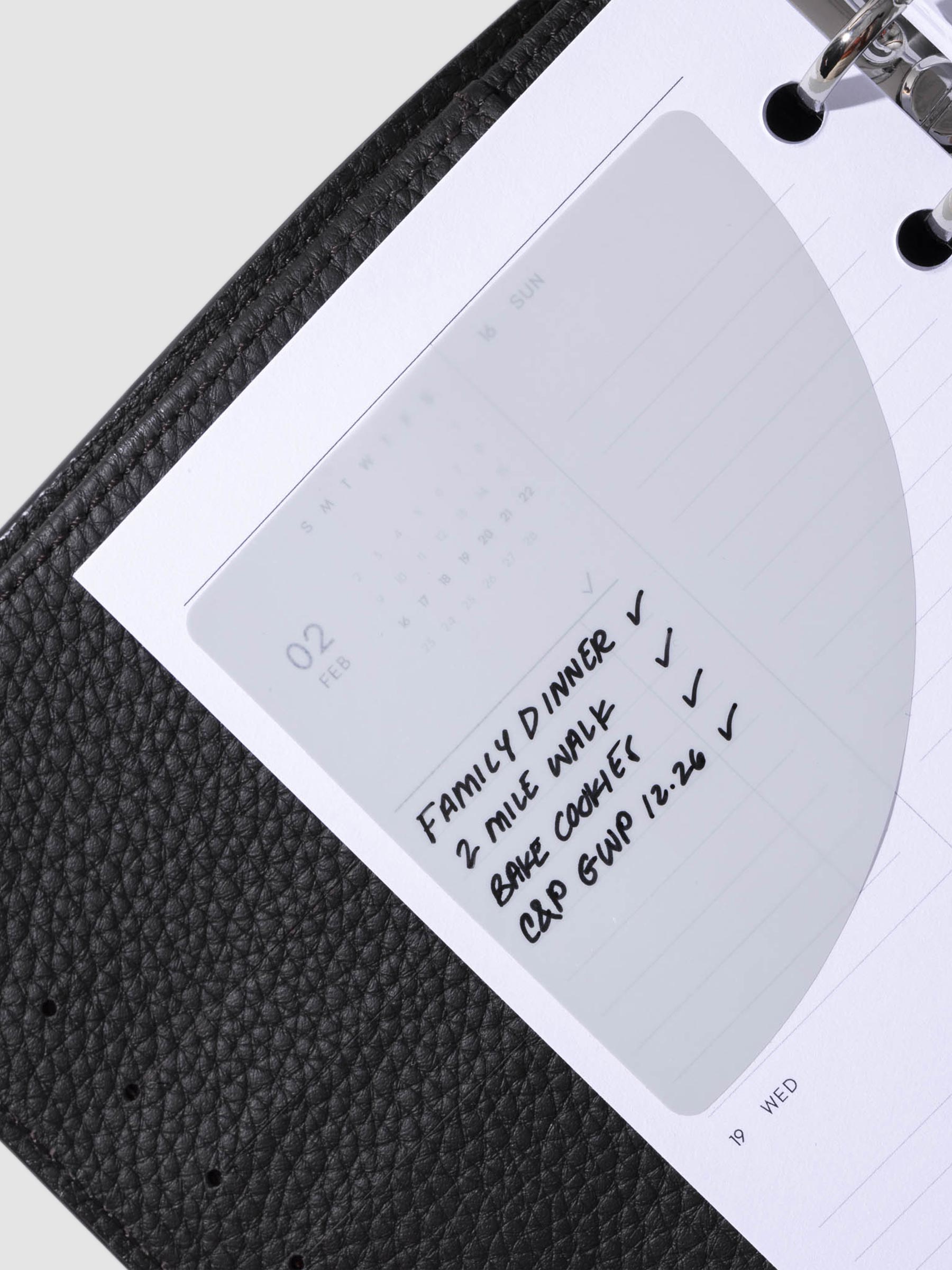 Sticky note in use inside a black leather planner. It is written on with a black oil based ink pen.