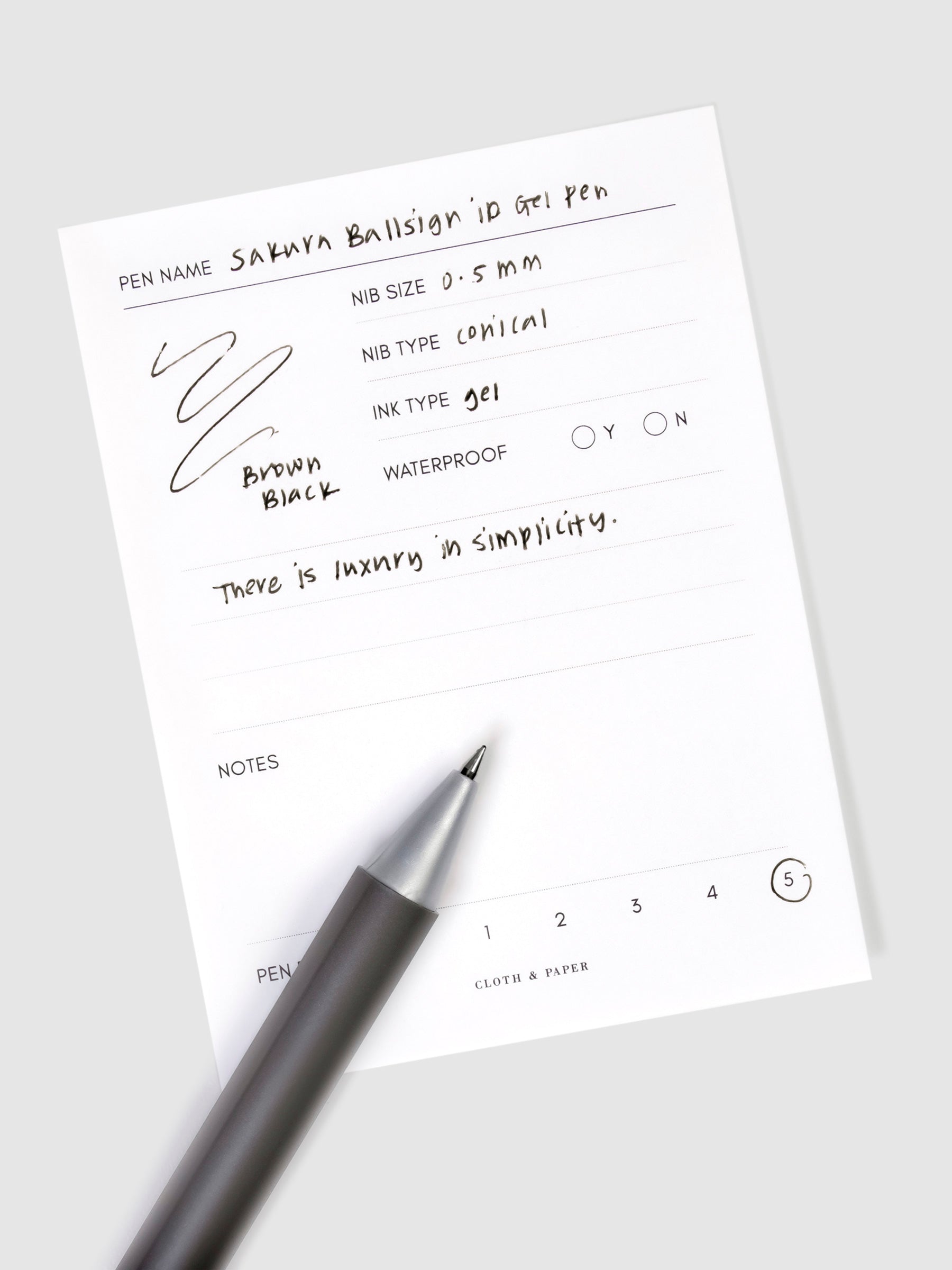 Pen in Brown Black resting on a pen test sheet displaying a writing sample detailing the pen's specs.