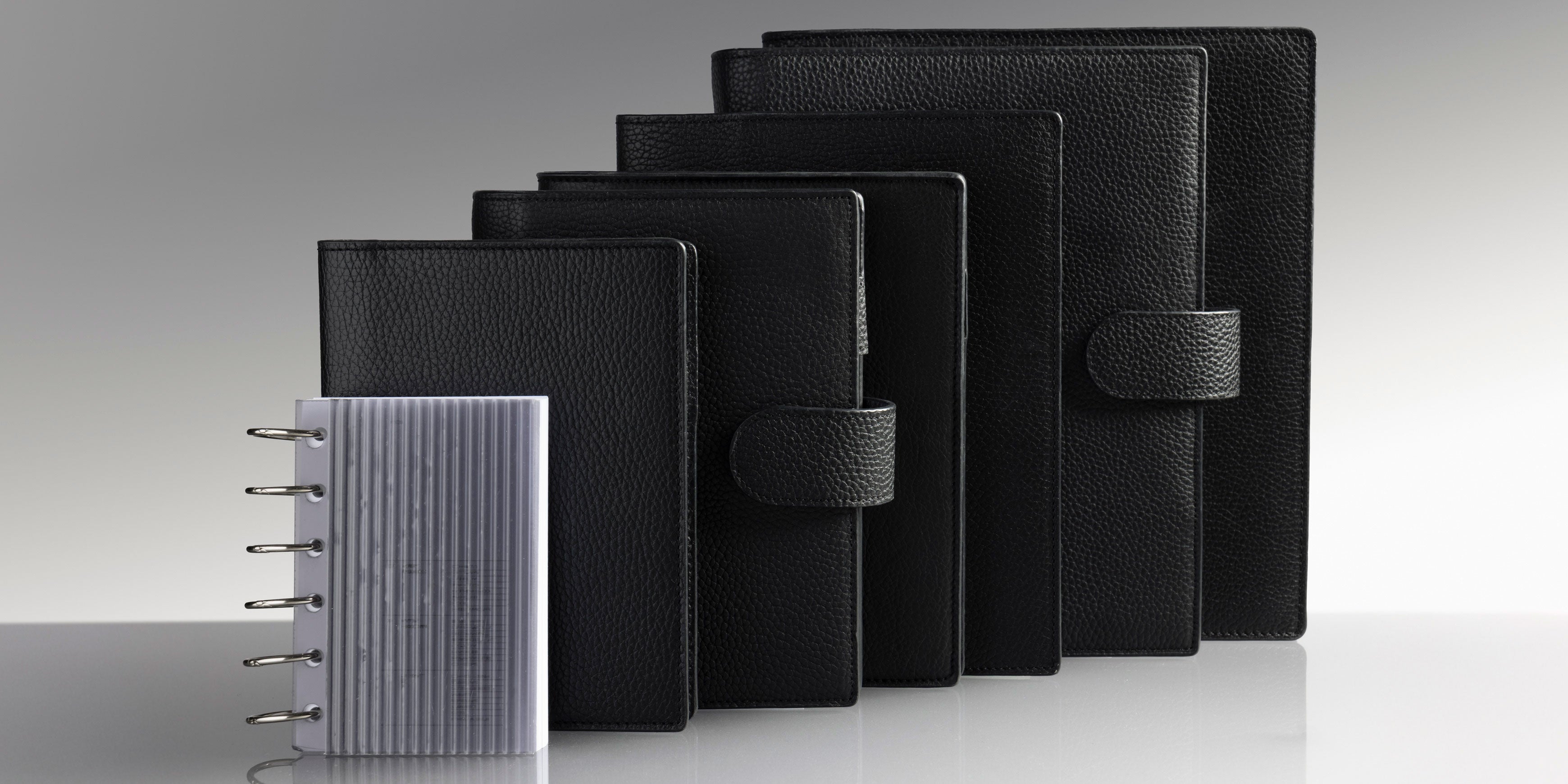 Lineup of black planner covers from small to large, with a small discbound insert on the left, on a gray reflective surface.