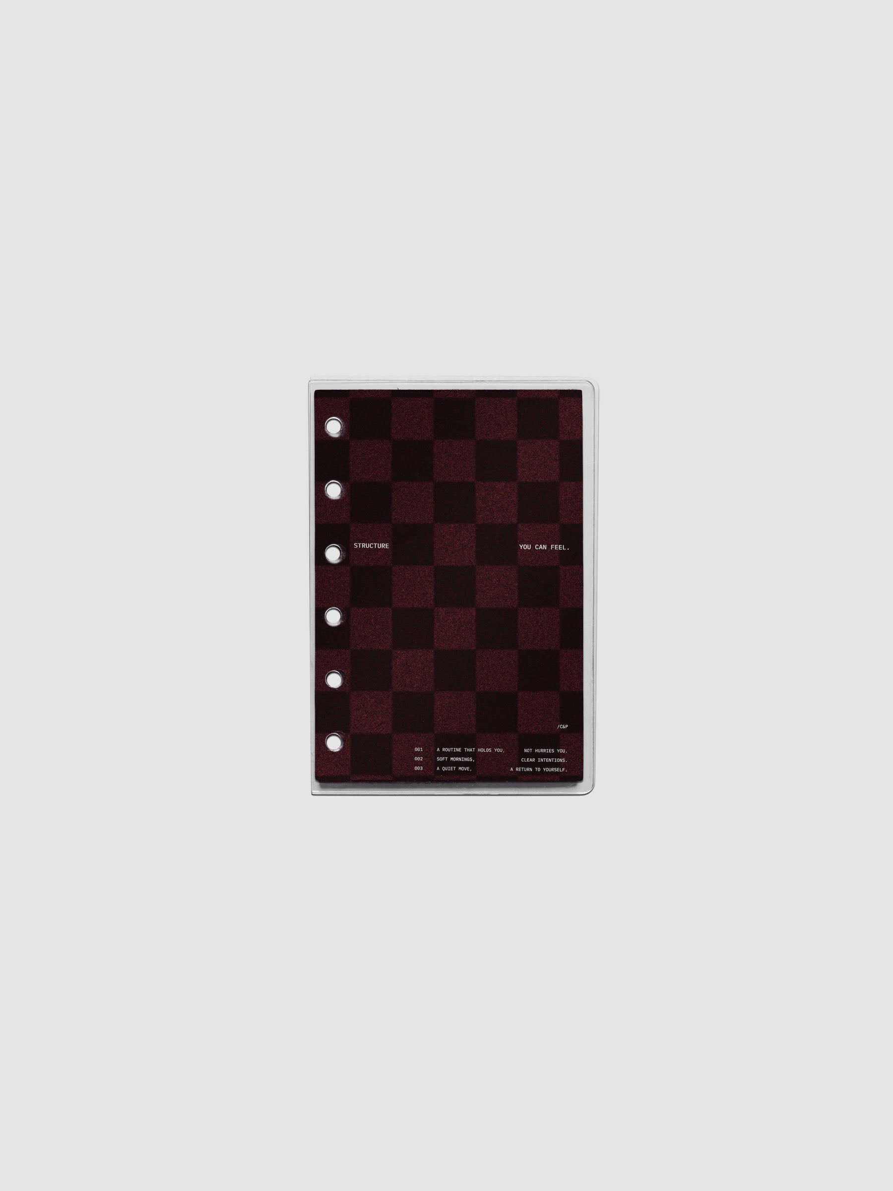 Pocket sized velvet checkered dashboard in the color Bordeaux in between two crystal clear plastic planner covers with text printed on the cover on the theme of structure and routines.