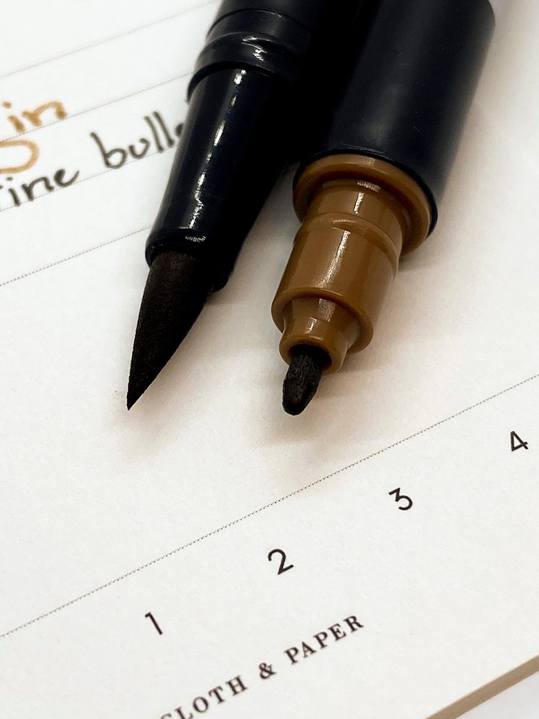 Tombow Dual Brush Art Marker, Saddle Brown, Cloth & Paper. Close up of the brush tip and fine tip of two Saddle Brown art markers resting on a pen test sheet.