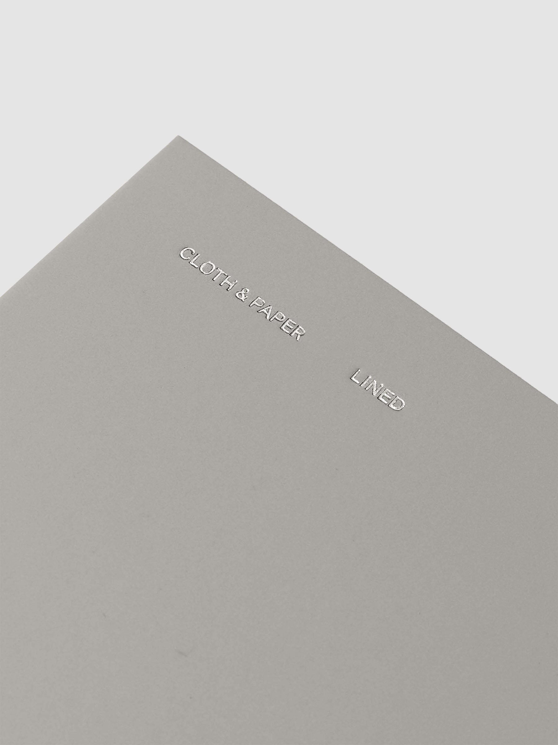 Travel Notebook in size Pocket Plus in the color Ash (grey) containing lined notes layouts shown closed from the front as a closeup on the foil details reading 'Cloth & Paper' 'Lined'.
