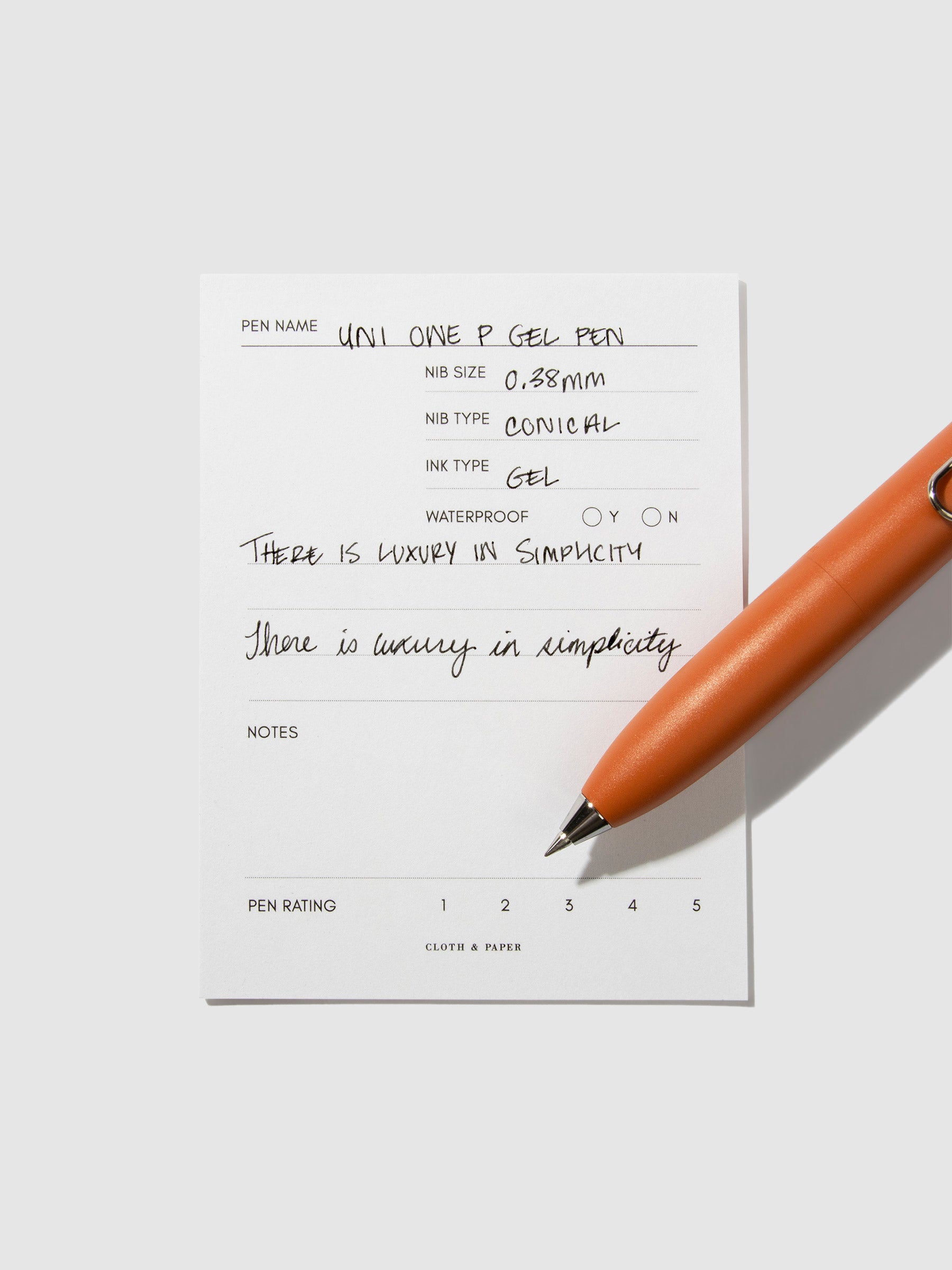 Uni-Ball One P pen resting on a pen test sheet. The sheet shows the pens specs. Color pictured is Mandarin Orange.