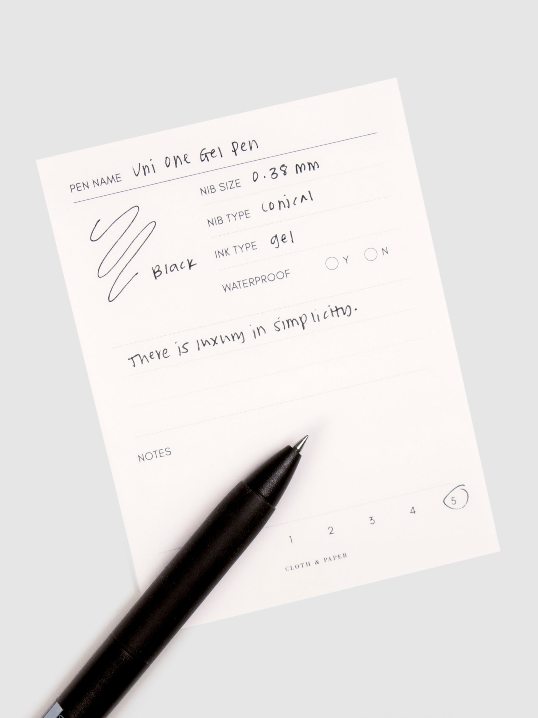 Uni One Gel Pen, 0.38 mm, Black Ink, Black Barrel, Cloth and Paper. Pen resting on pen test sheet displaying writing sample.