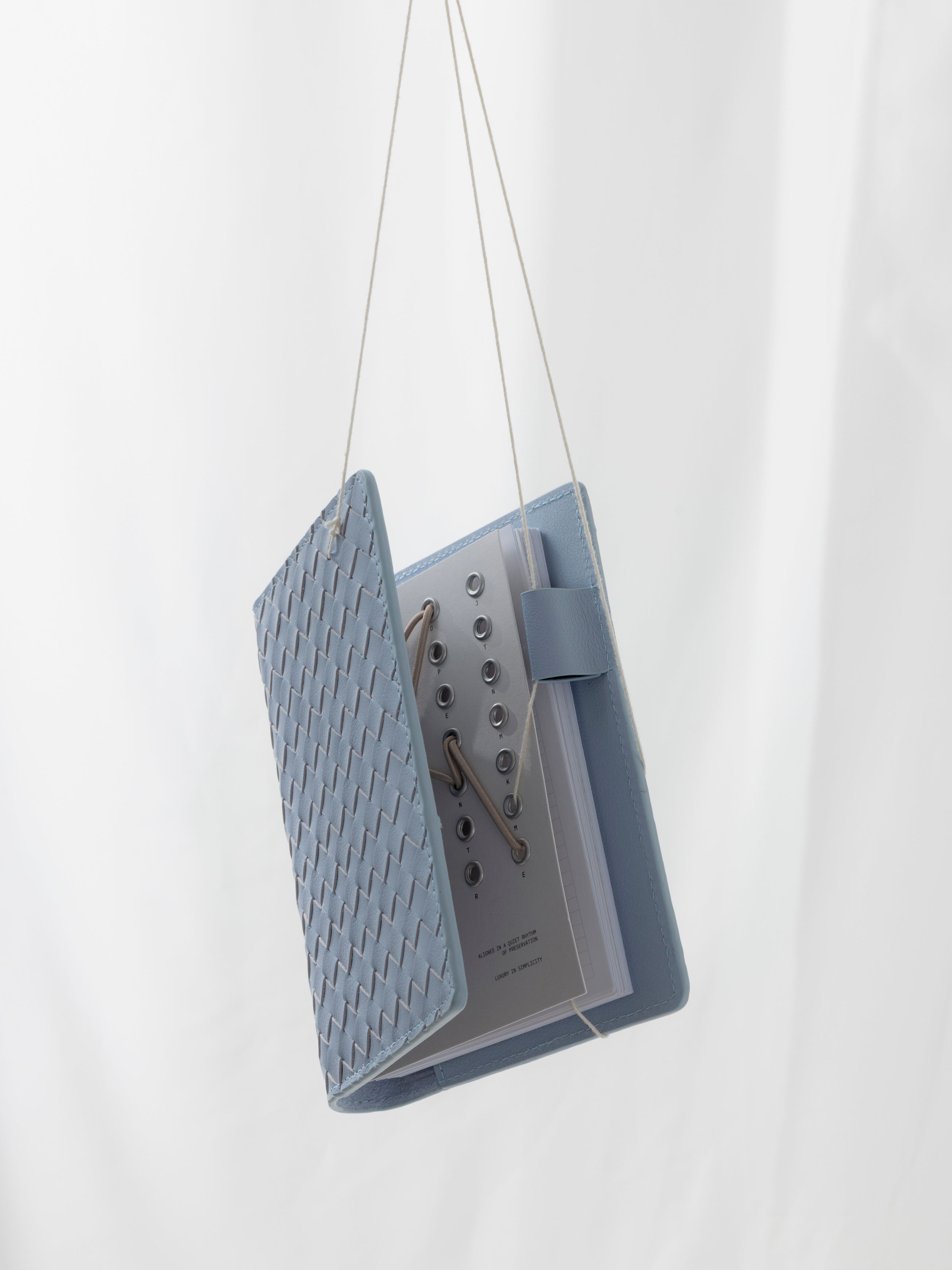 A light blue woven leather planner cover hangs from string against a light, fabric background.