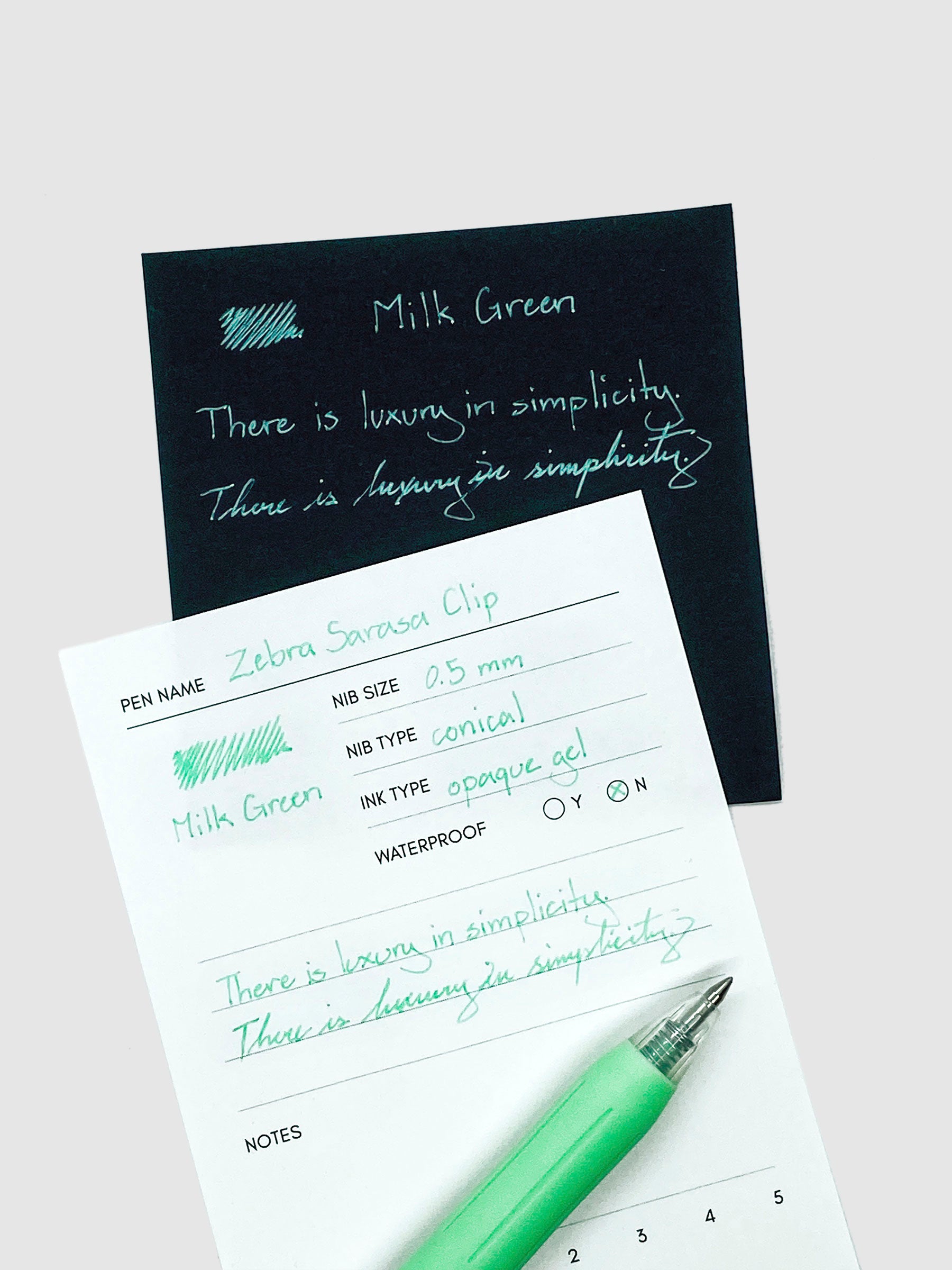 Zebra Sarasa Clip, 0.5 mm, Milk Green, Cloth and Paper. Pen resting on pen test sheet displaying writing sample with black paper behind it showing alternate writing sample.