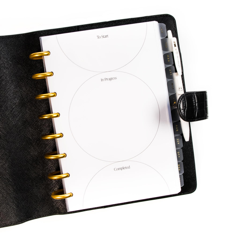 Kanban Planner Dashboard styled in a discbound planner tucked into a black leather planner cover. A white pen is inside the planner cover's pen loop.