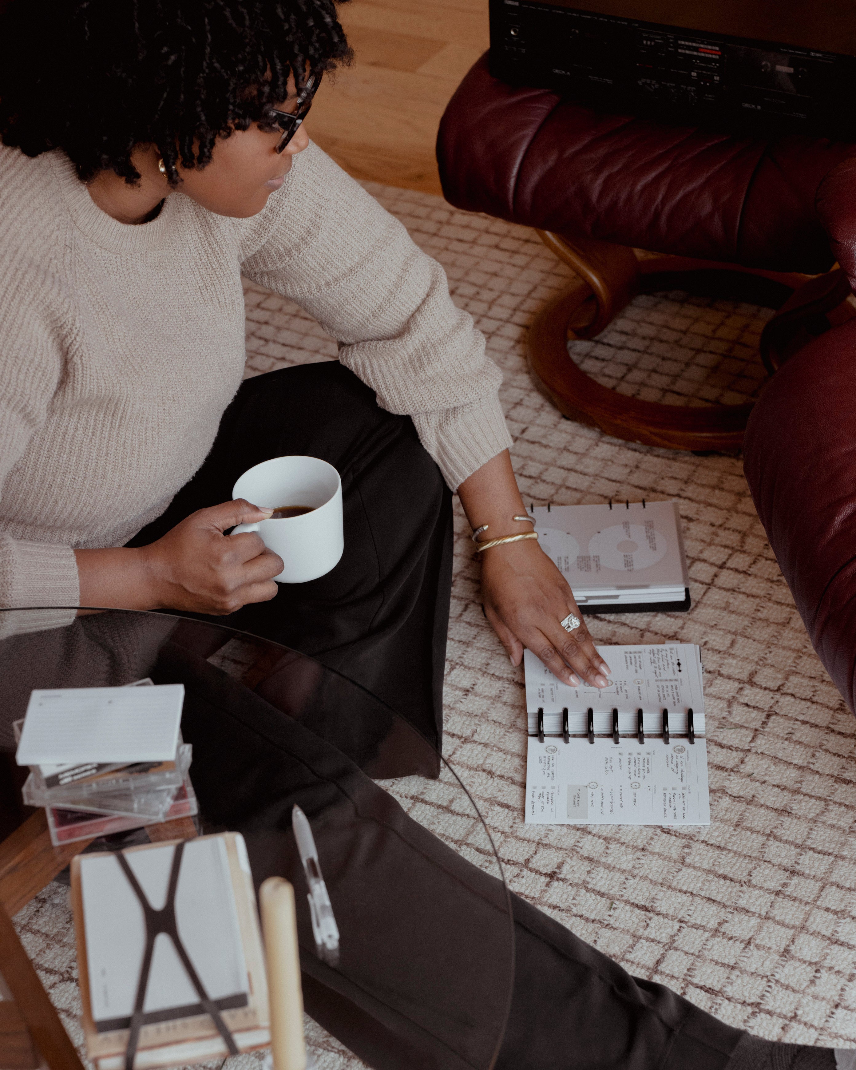A person sitting on a carpeted floor, holding a white mug and looking down at an open disc-bound planner surrounded by February 2026 Intention Box items.