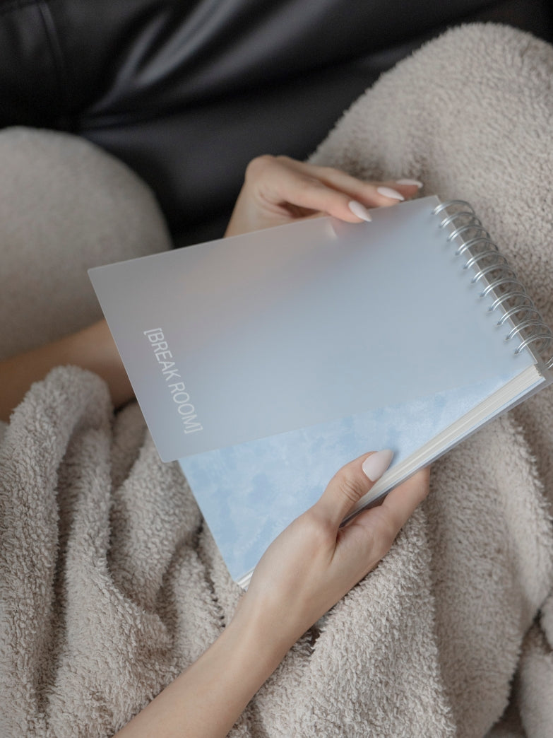 Person sitting on a black leather couch wrapped in a beige fleece blanket, holding an open Break Room Spiral-bound Notebook.