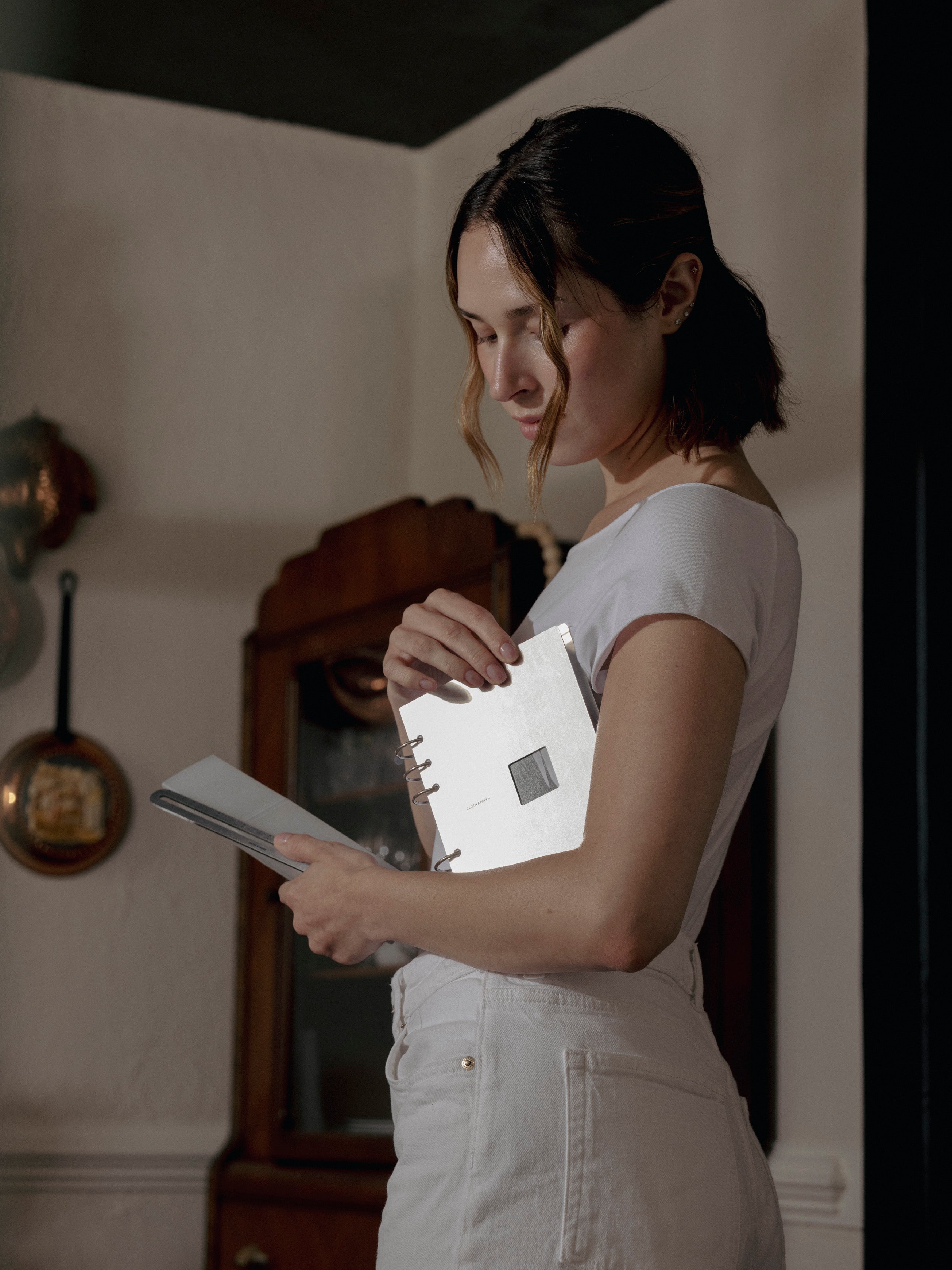 Woman holding a silver planner standing in a room with a wooden cabinet and decorative items.