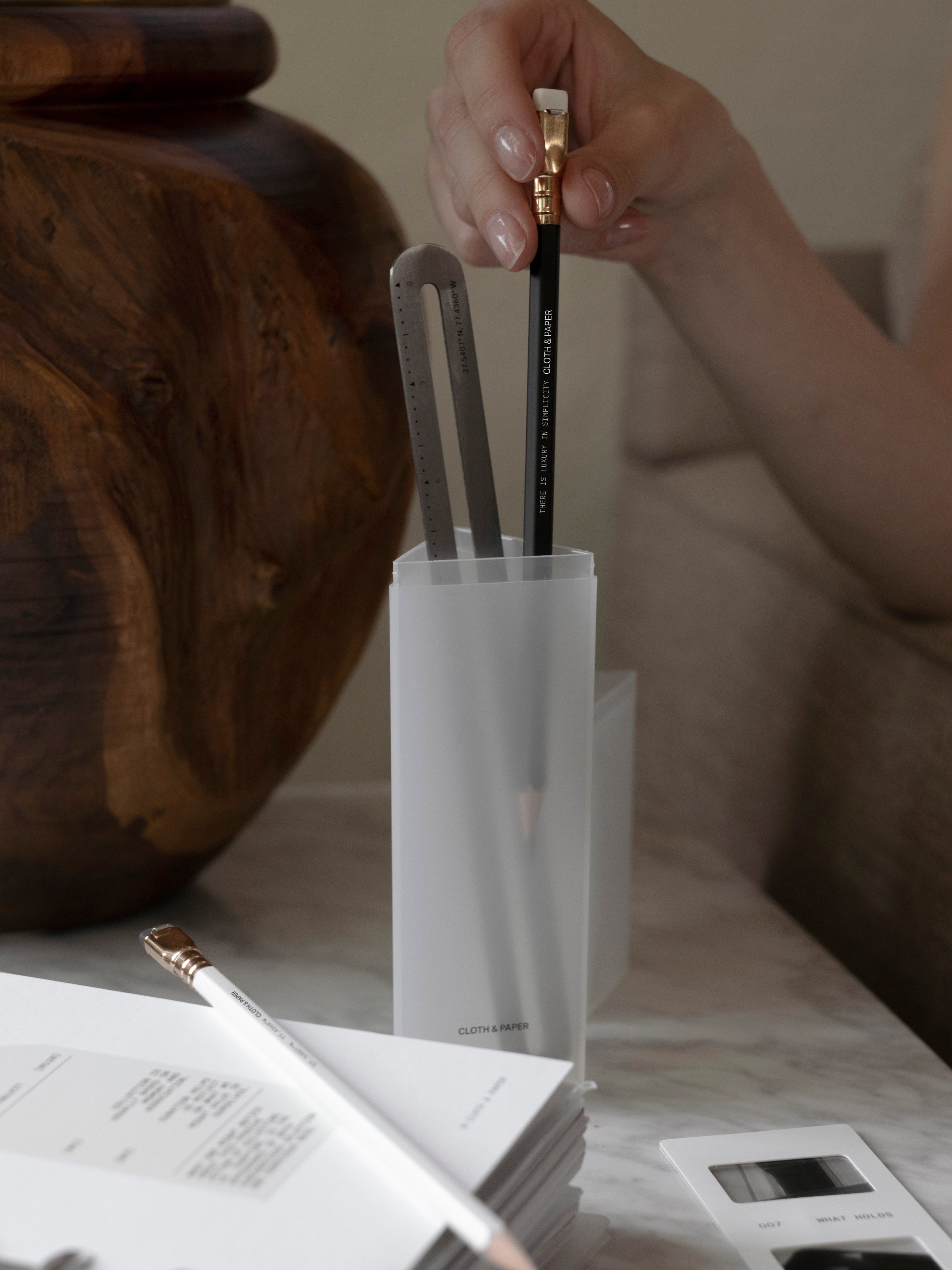 Hand removing a black pencil from a frosted Pencil Box standing upright on a marble surface, with a stainless steel ruler, white pencil, and open planner insert visible in the foreground