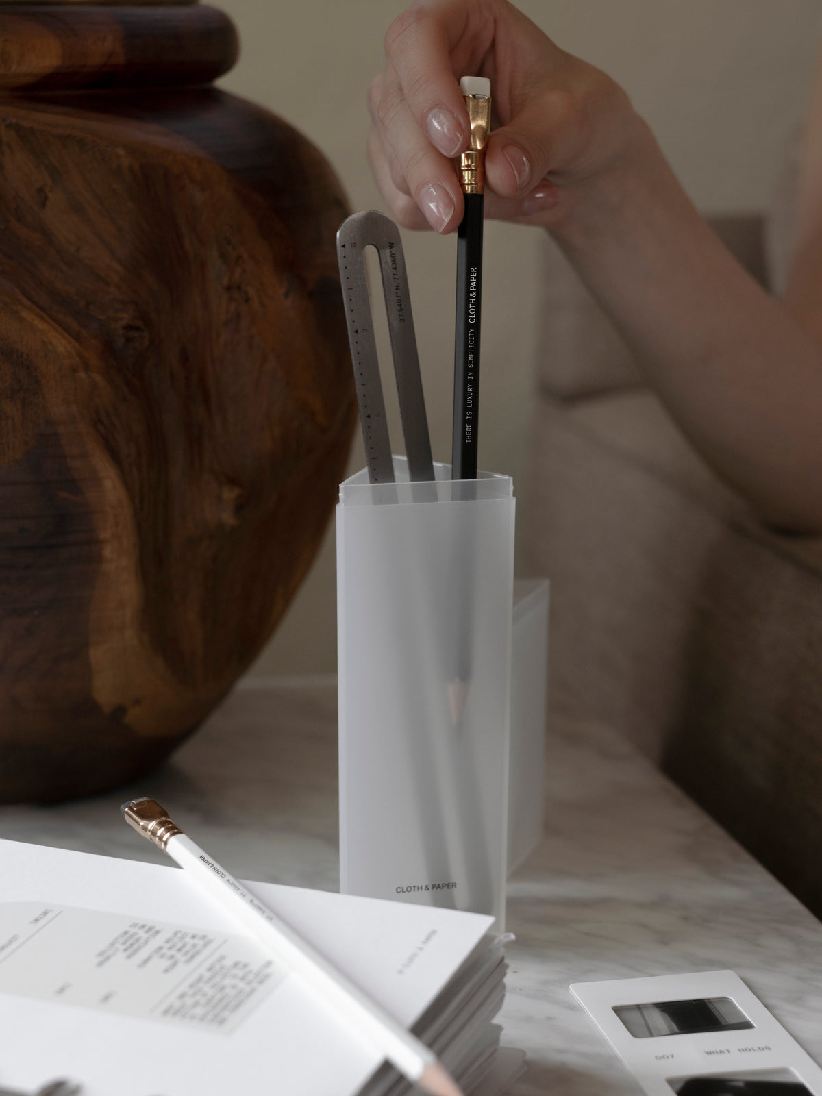 Hand removing a black pencil from a frosted Pencil Box standing upright on a marble surface, with a stainless steel ruler, white pencil, and open planner insert visible in the foreground