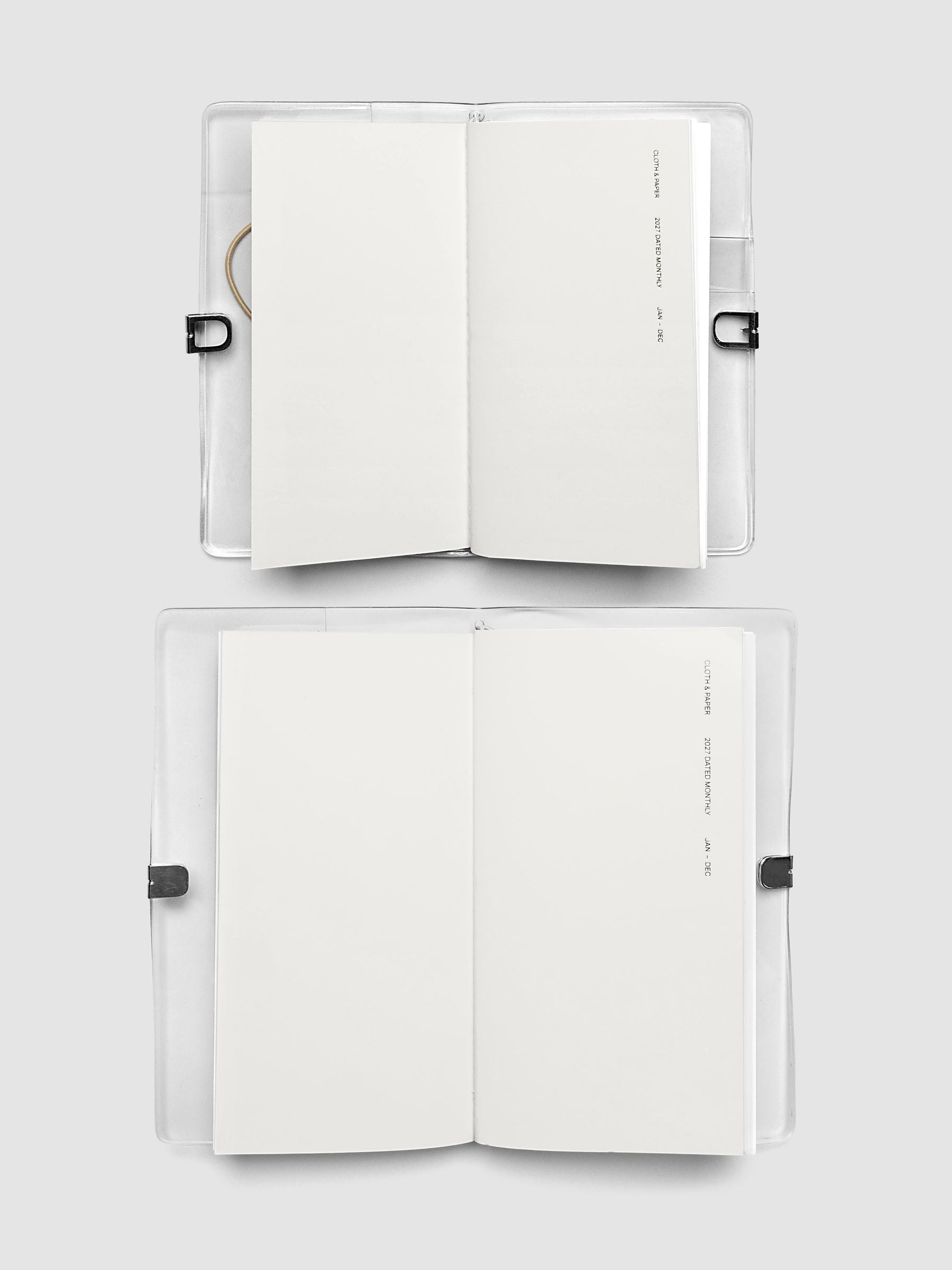 Two open travel notebooks stacked in their clear vinyl covers, both showing a blank interior page and 2027 Dated Monthly January through December cover text, comparing Pocket Plus and A5 Slim sizes