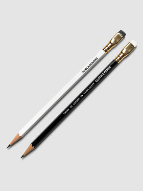 Two pencils, one white and one black, both with gold ferrules and erasers, laid diagonally on a light gray background. The white pencil is branded 'BLACKWING', and the black pencil reads 'THERE IS LUXURY IN SIMPLICITY' with the Cloth and Paper logo. 