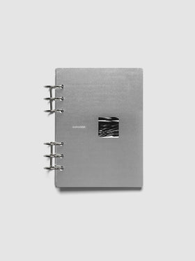 A silver-gray 6-ring-bound notebook with a small black-and-white abstract square inset on the front cover, photographed against a light gray background.