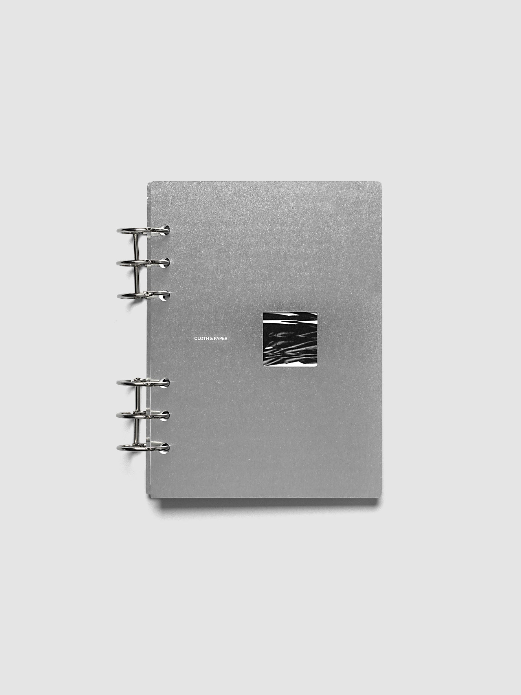 A silver-gray 6-ring-bound notebook with a small black-and-white abstract square inset on the front cover, photographed against a light gray background.