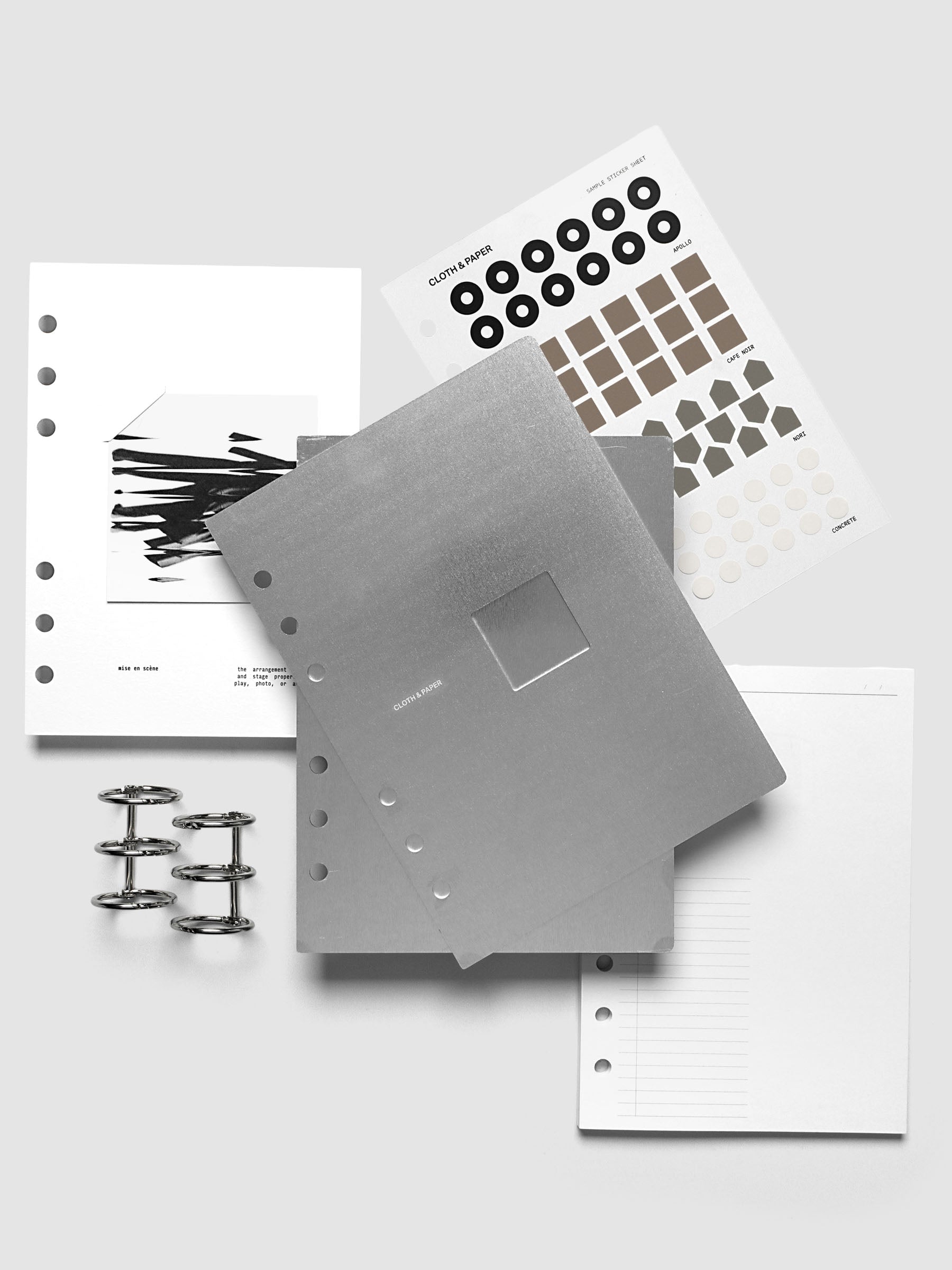 A flat-lay of a metal cover and planner bundle set on a light gray background. The set includes a brushed silver metal binder cover with a square window, planner inserts, a sticker sheet and silver metal binder rings.