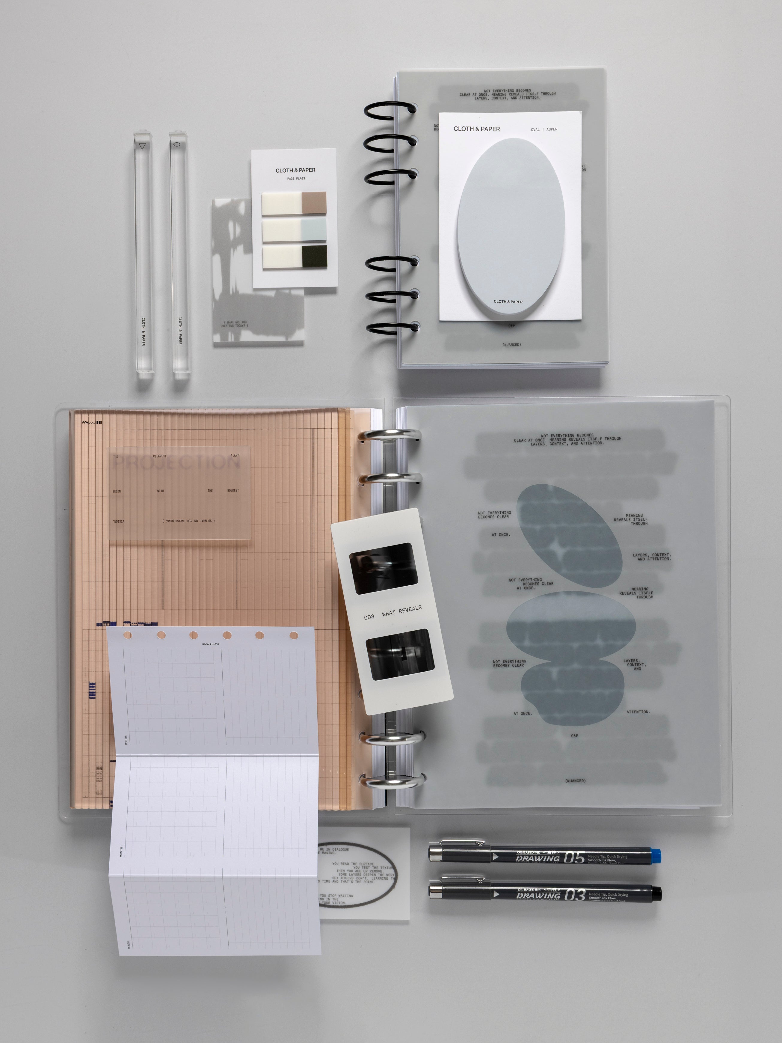 August Intention Box contents arranged on a light gray surface, including two clear pen cases, a page flag set, transparent sticky notes, an oval sticky note pad, a ring bound planner open to an amber corrugated insert and a frosted printed dashboard, a notepad, and two black drawing pens