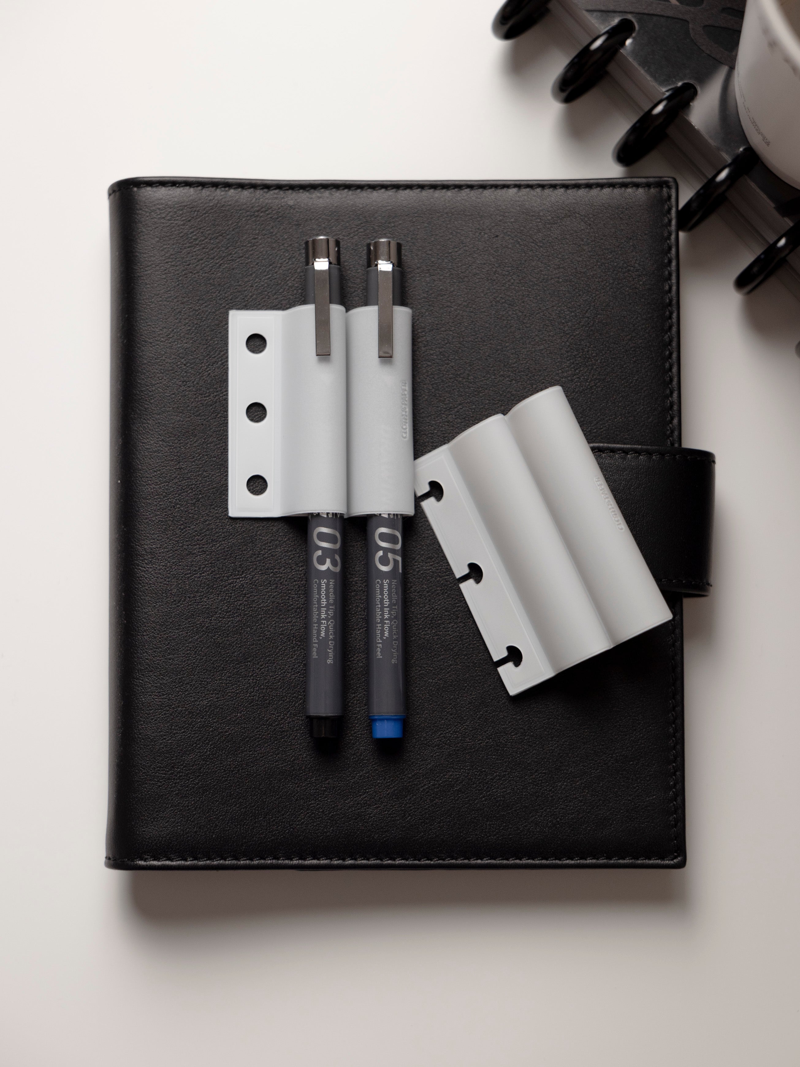 Black leather planner cover shown with two gray pen sleeves, three hole punched, holding two black drawing pens