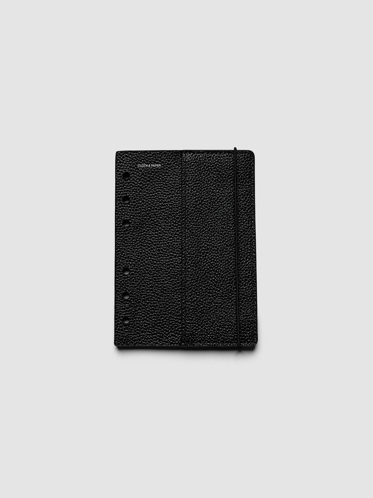 The front cover of the A6 Genuine Leather Planner Cover Set laid flat, showing pebbled black leather, the silver foiled logo at the top left, the punch holes down the left edge, and the bungee cord running down the right