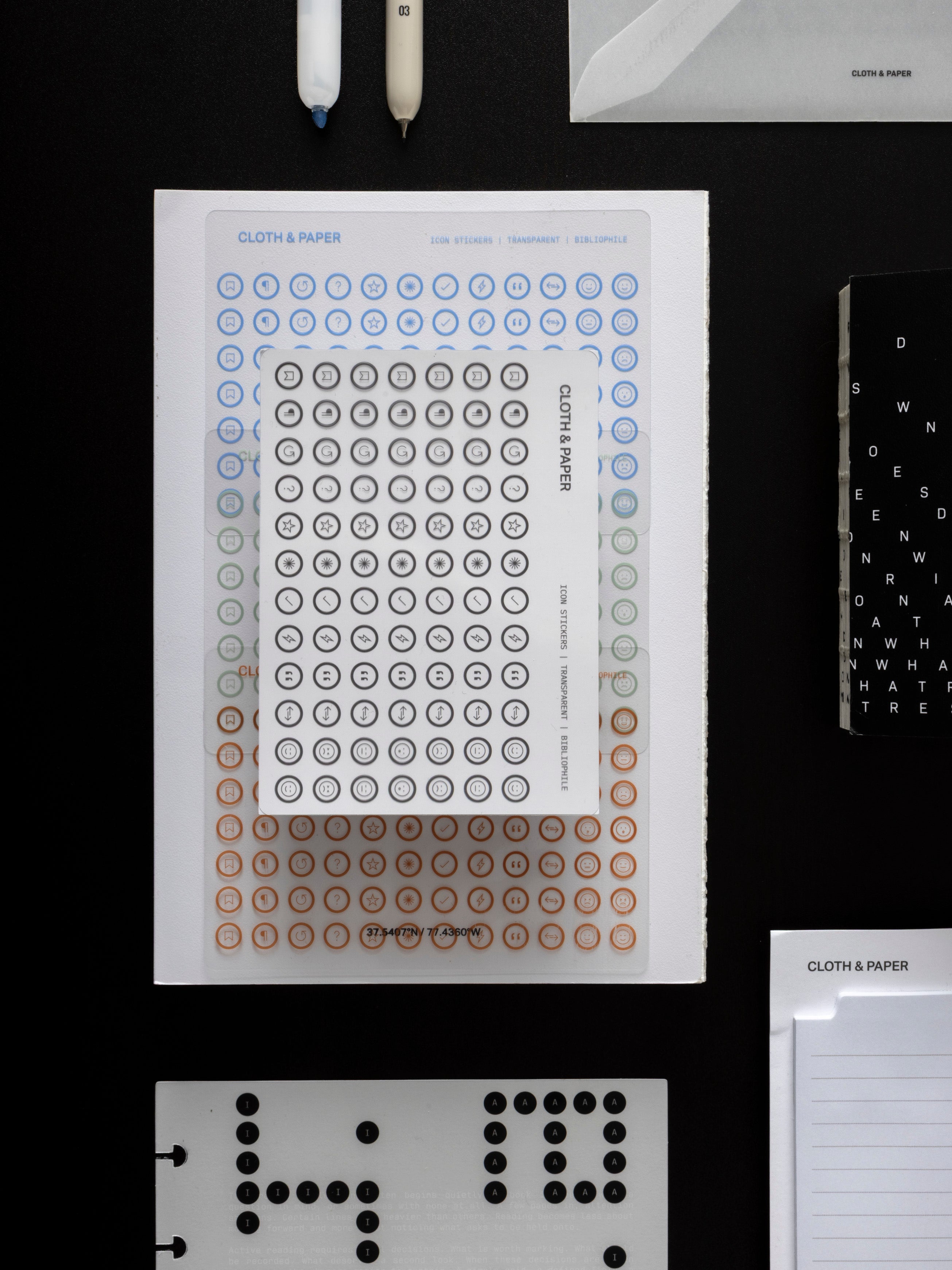Icon Stickers sheets layered on a black surface, with a highlighter and a fine liner pen above, the Bibliophile Notebook to the right, and a Scantron dashboard below.