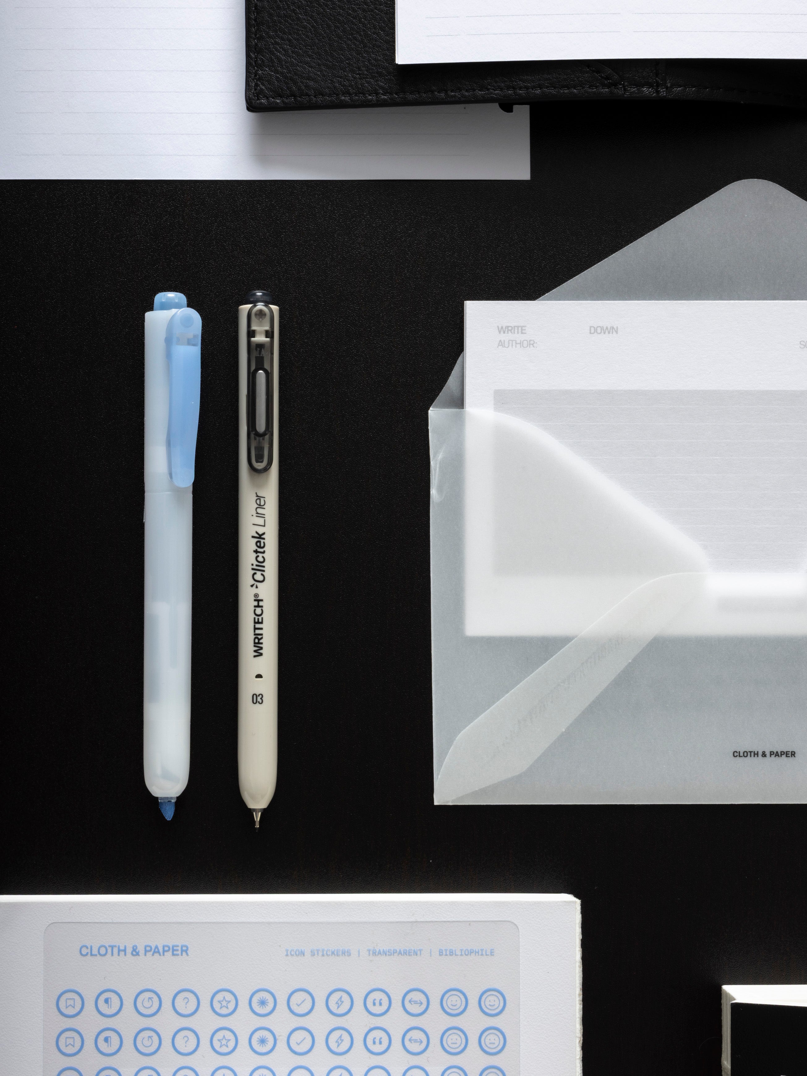 A highlighter and a fine liner pen laid side by side on a black surface, beside a vellum envelope holding an Archive Index Card, with an Icon Stickers sheet below.